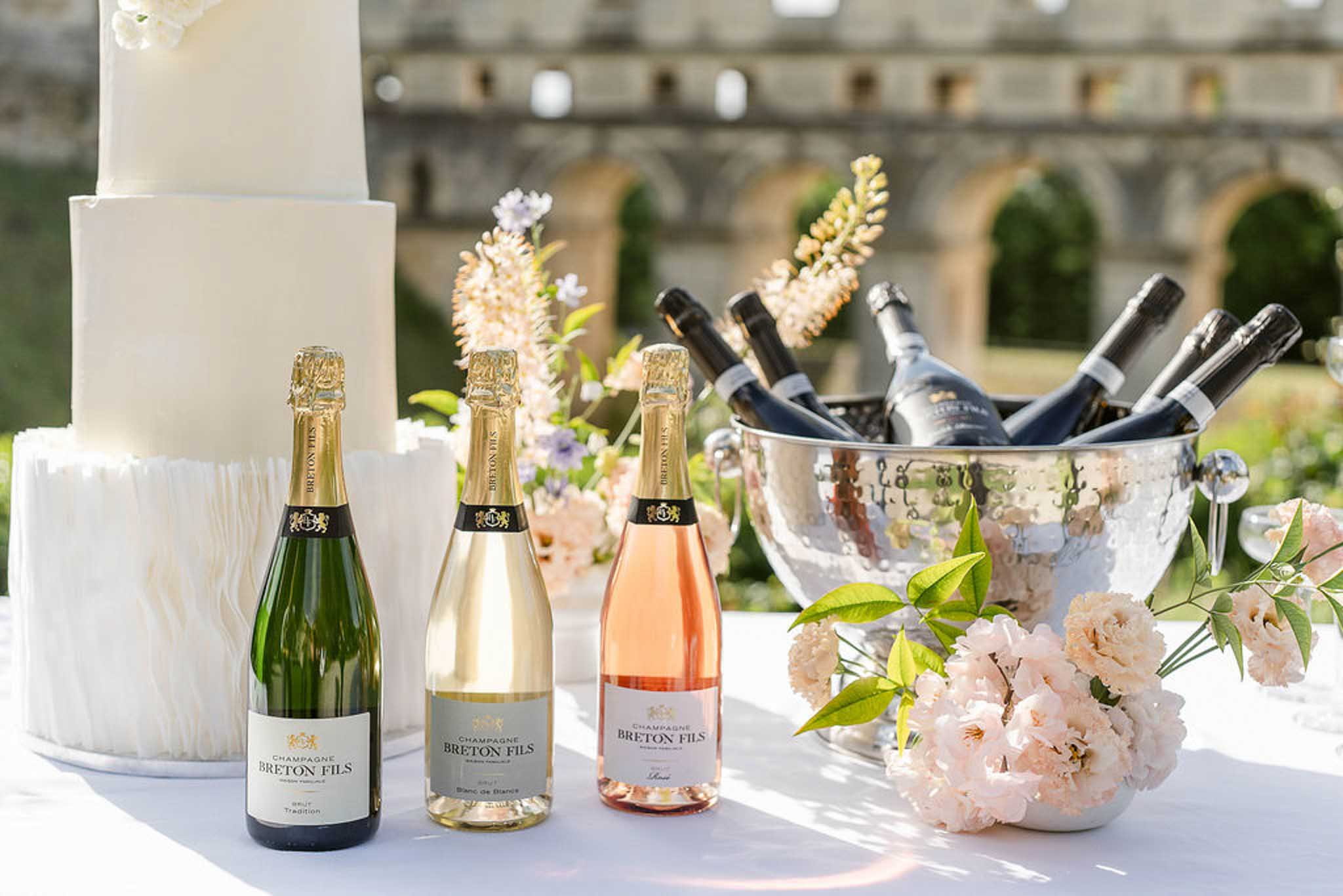 White cake beside Breton Fils champagne bottles and blush peony arrangement with silver ice bucket in courtyard