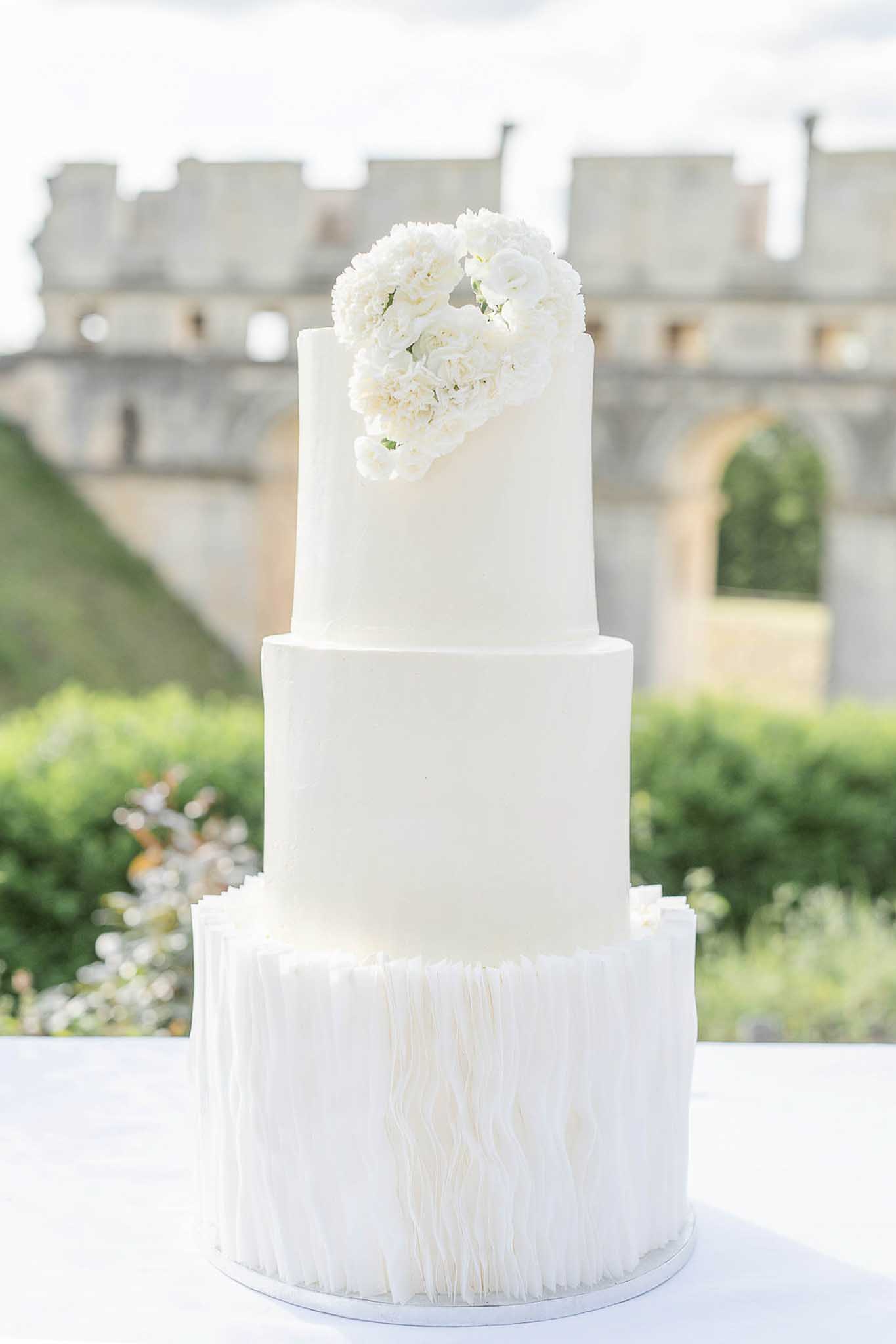 Three-tiered white wedding cake topped with white peonies and garden roses displayed in a chateau courtyard