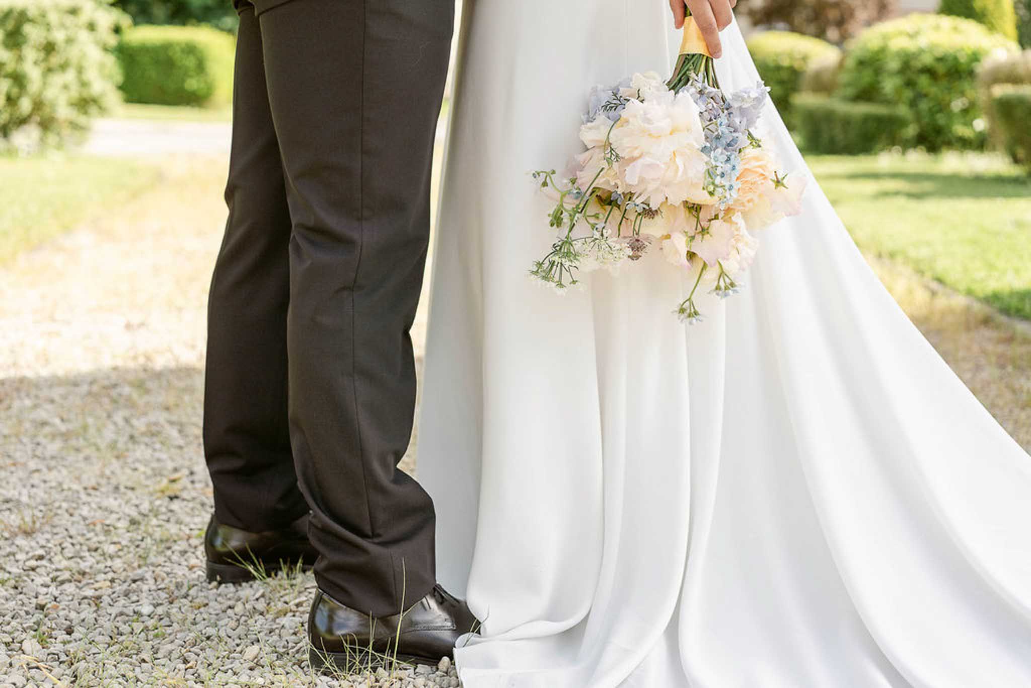 Close-up of bridal bouquet of white peonies, peach roses and blue-grey hydrangeas held by bride in ivory dress on gravel path