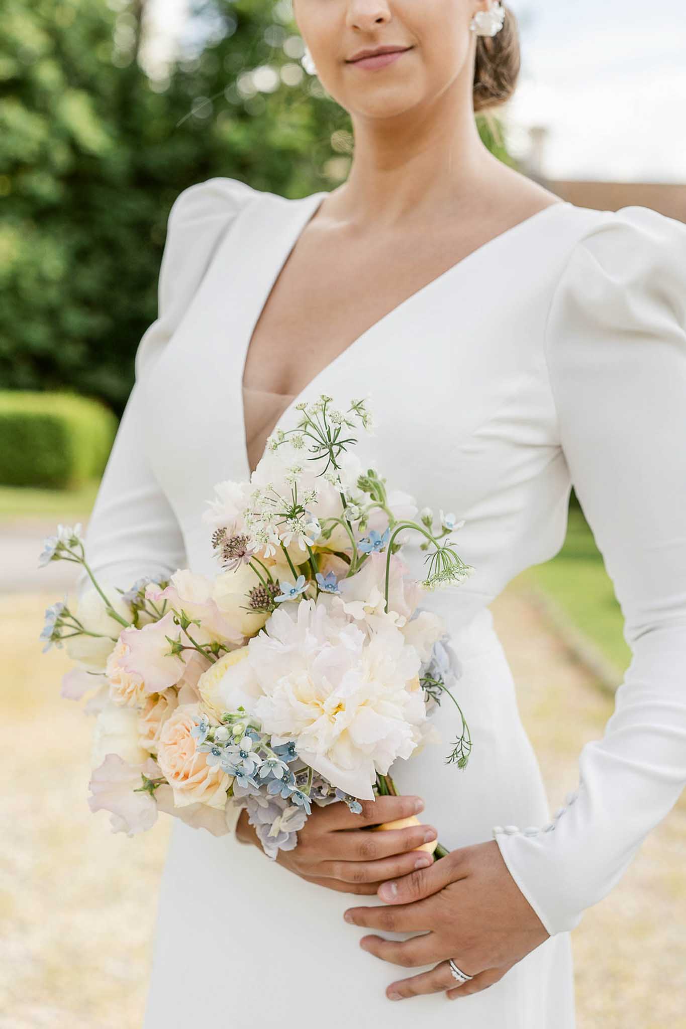 Bride in V-neck dress with puffed sleeves holding bouquet of peach peonies, blue hydrangeas, and white astilbe