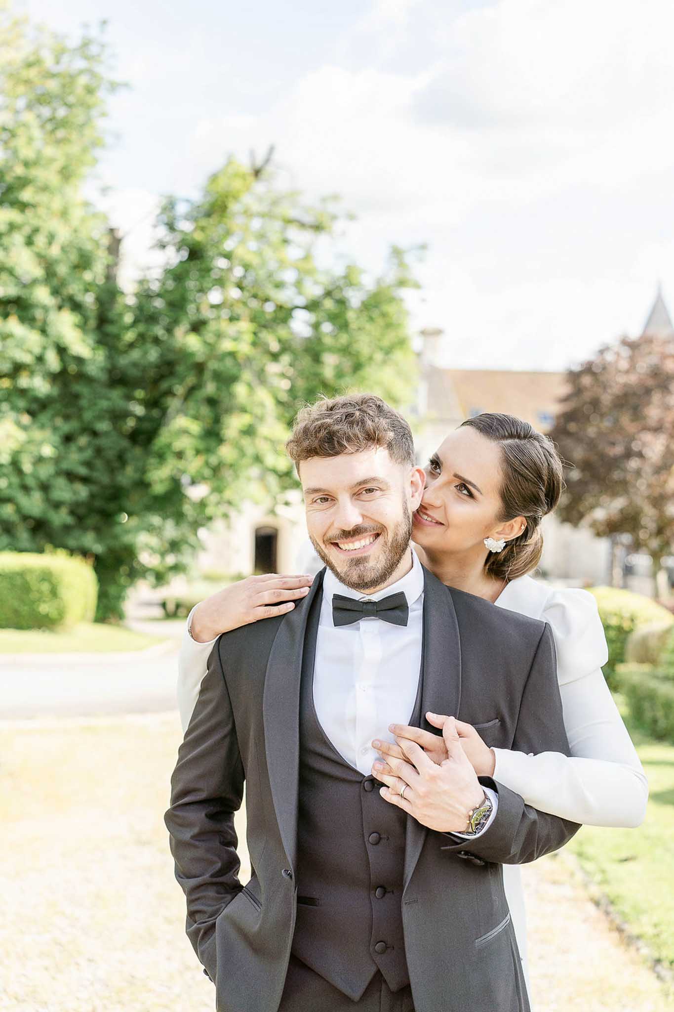 Newlywed couple smiling in estate garden, groom in charcoal tuxedo and bride in ivory sleeveless dress
