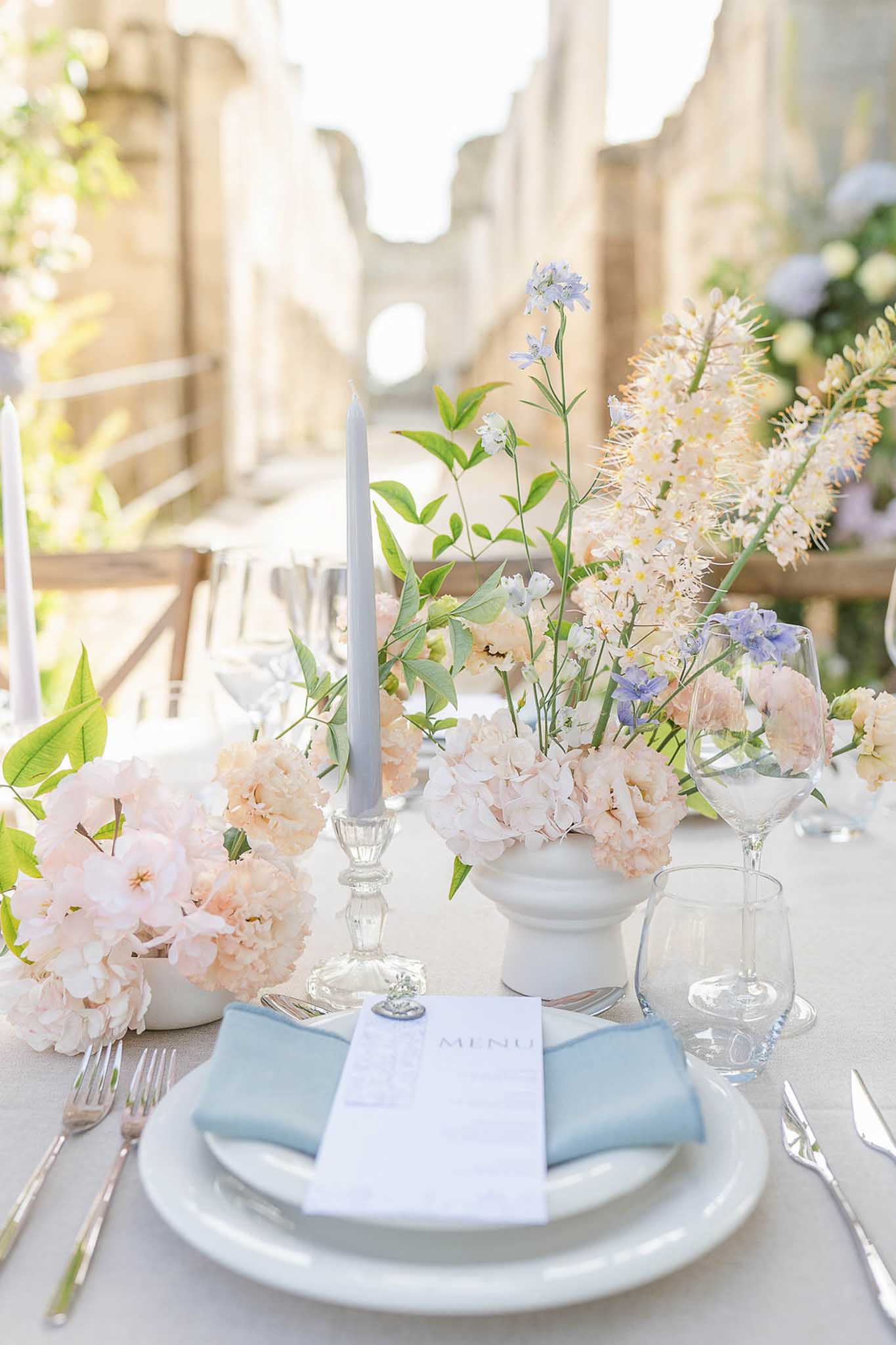 Reception place setting with blue napkin, white menu card, and blush hydrangea centerpiece in stone courtyard