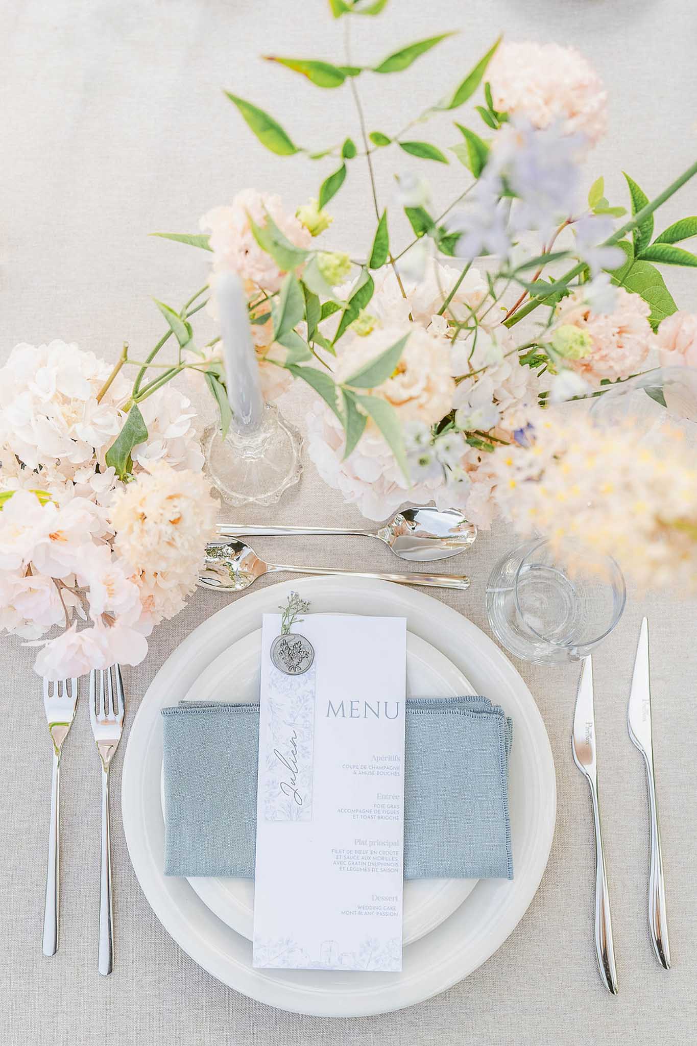 Overhead flat-lay of place setting with sage linen napkin, white plates, silver flatware, and ivory peonies with pale blue delphinium