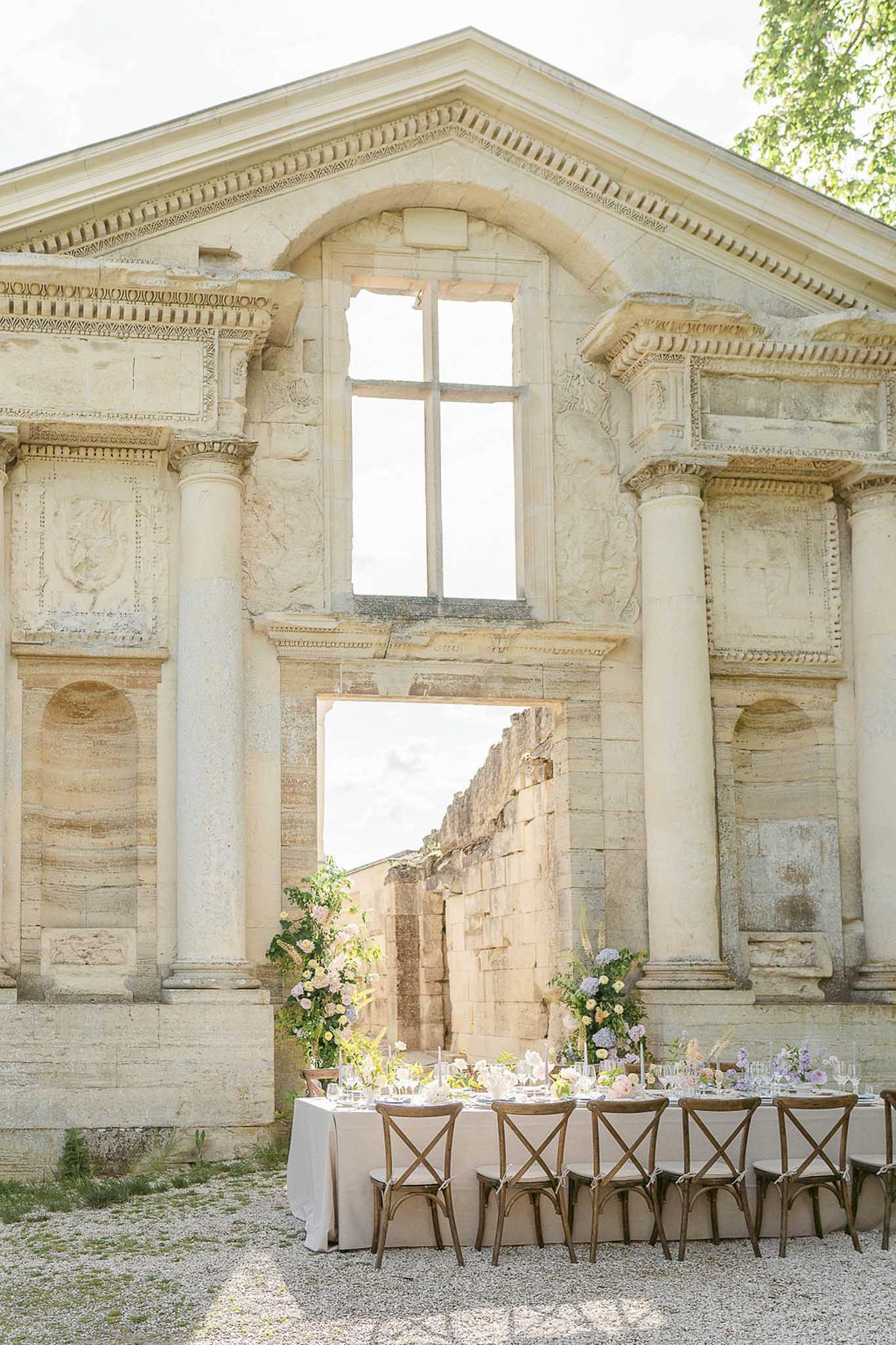 Outdoor reception table with cream linen and crossback chairs before stone classical building with lavender florals