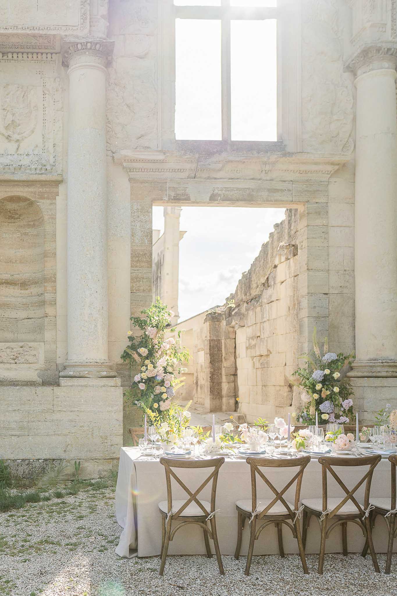 Long reception table set with ivory linens and floral arrangements inside stone ruins with Corinthian columns