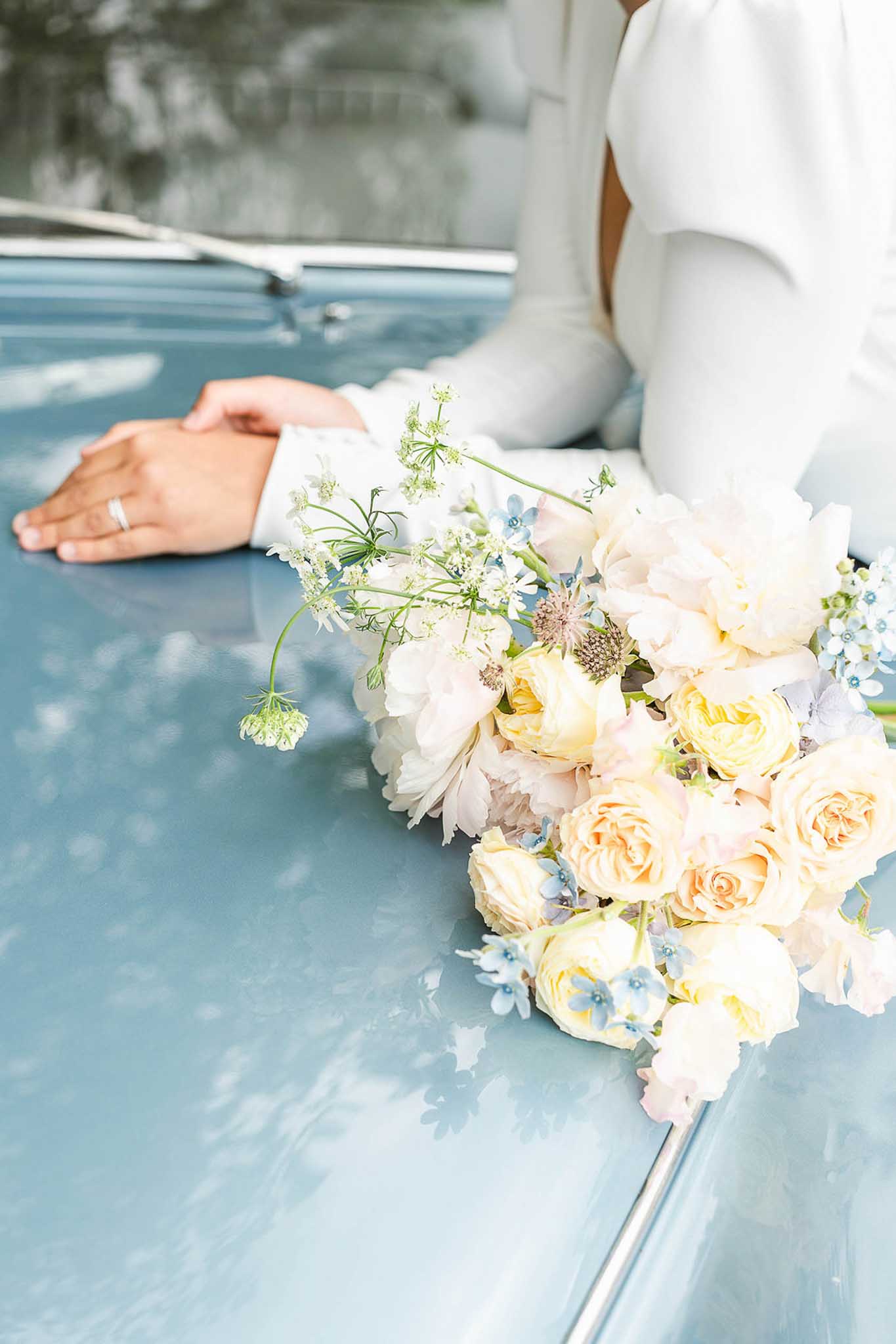 Close-up of bride's hands and garden-style bouquet of cream roses, white peonies and forget-me-nots resting on a pale blue vintage car.