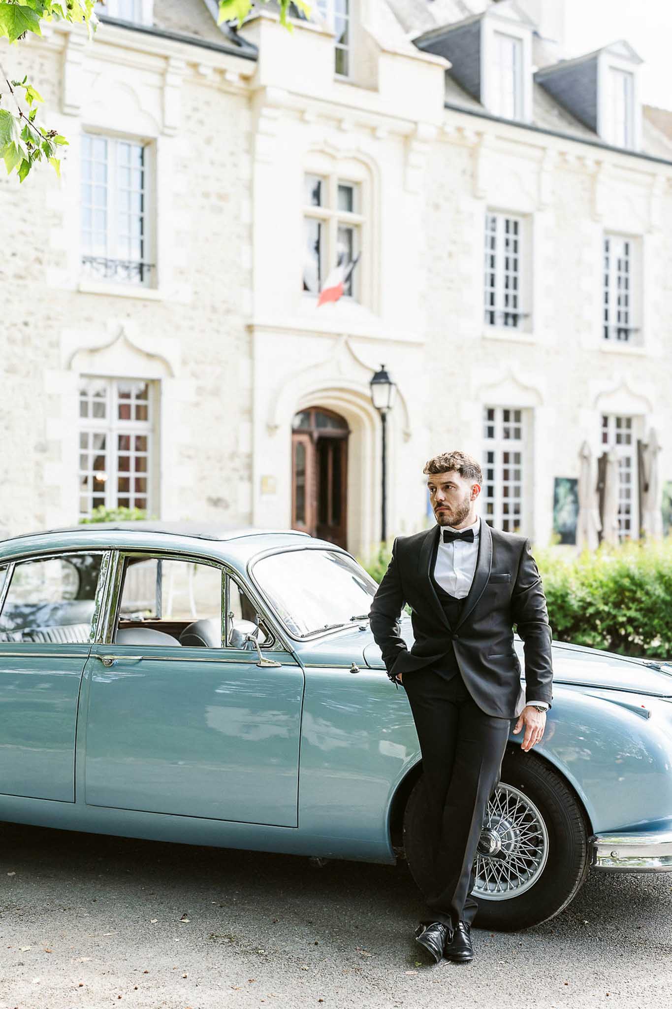 Groom in black tuxedo posing beside a vintage pale blue car in front of a cream stone manor house