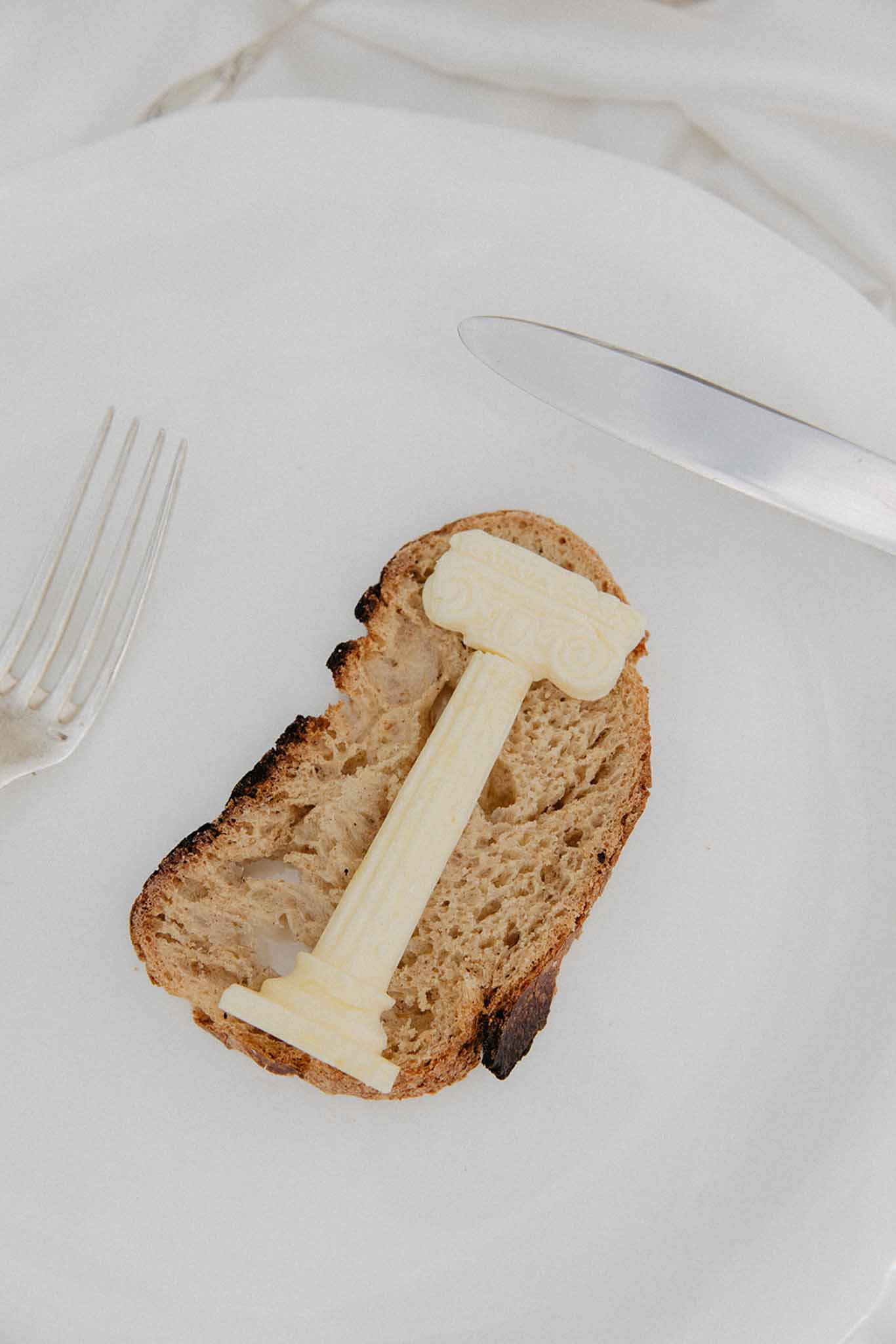 Minimalist plate with two butter pats on whole grain toast, fork and knife on white linen at reception dining service