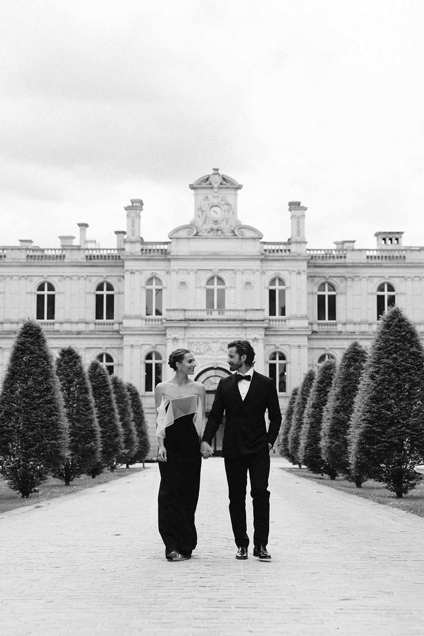 Bride and groom walking hand-in-hand through formal courtyard with cypress topiaries and neoclassical stone building behind
