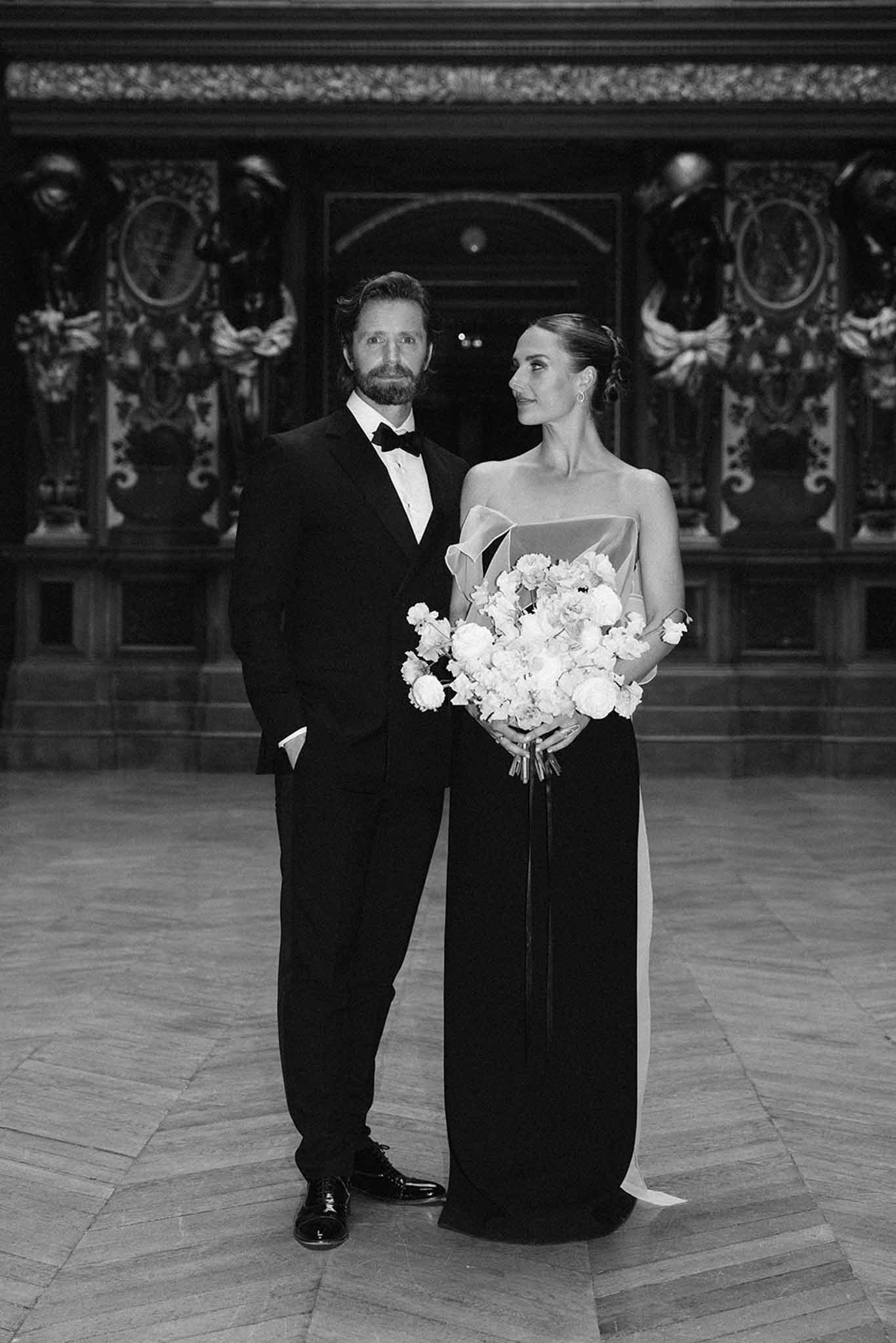 Bride in black strapless gown with white bouquet and groom in tuxedo in ornate classical interior, black and white