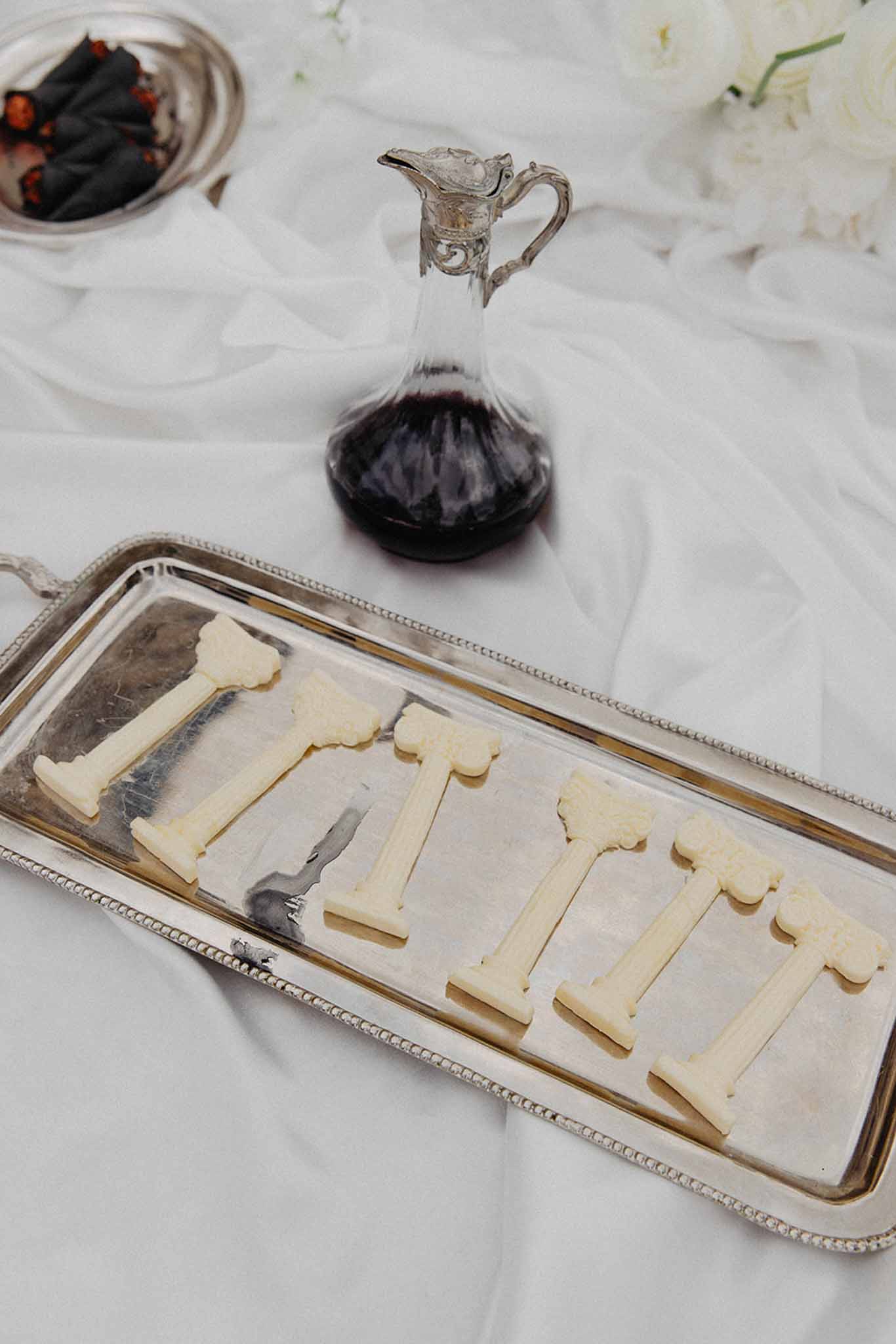 Close-up of silver serving pieces with butter servings, red wine decanter, and nuts on a white damask tablecloth