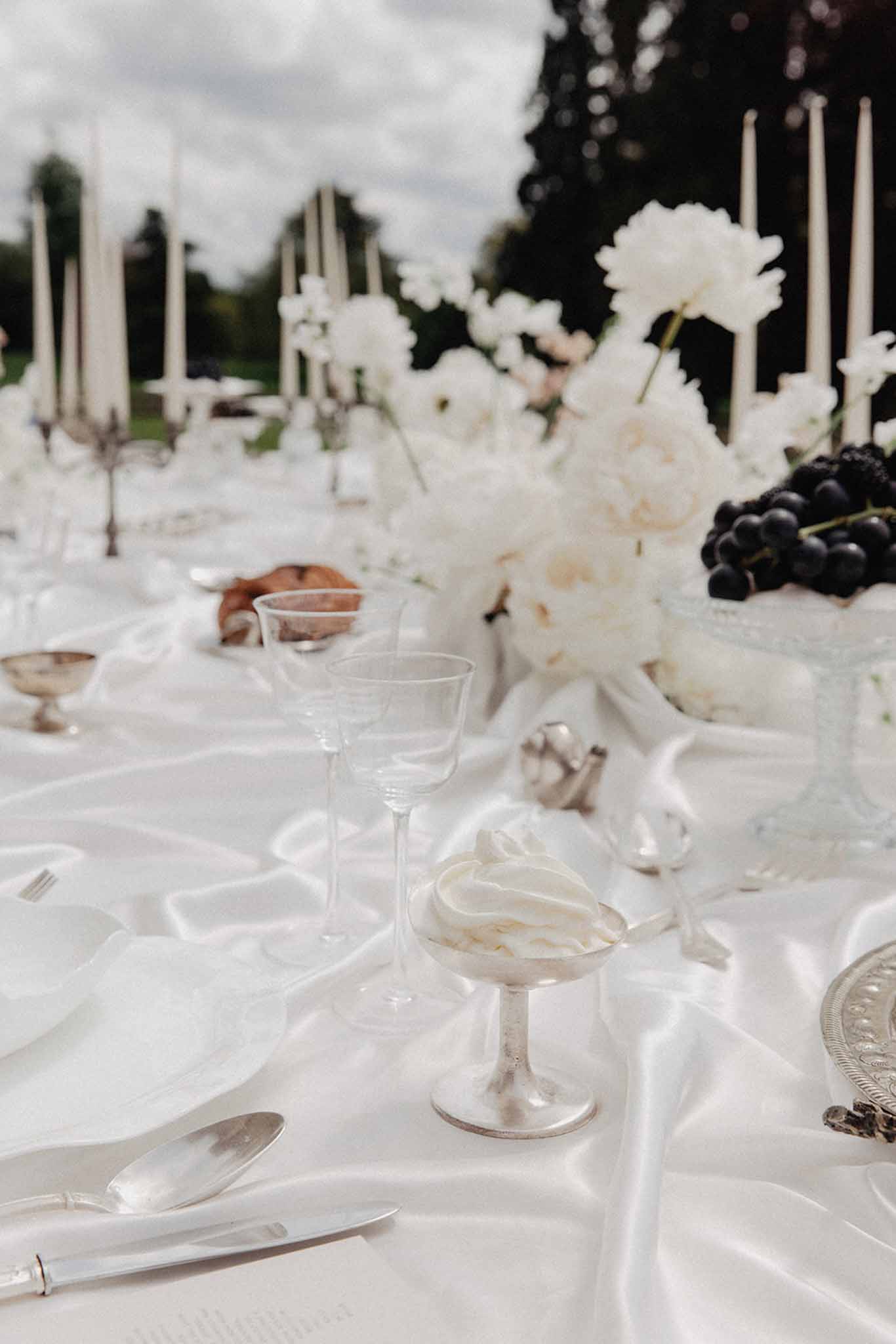 Outdoor reception table with white peony and rose centerpieces, ivory taper candles, and dark grape accents