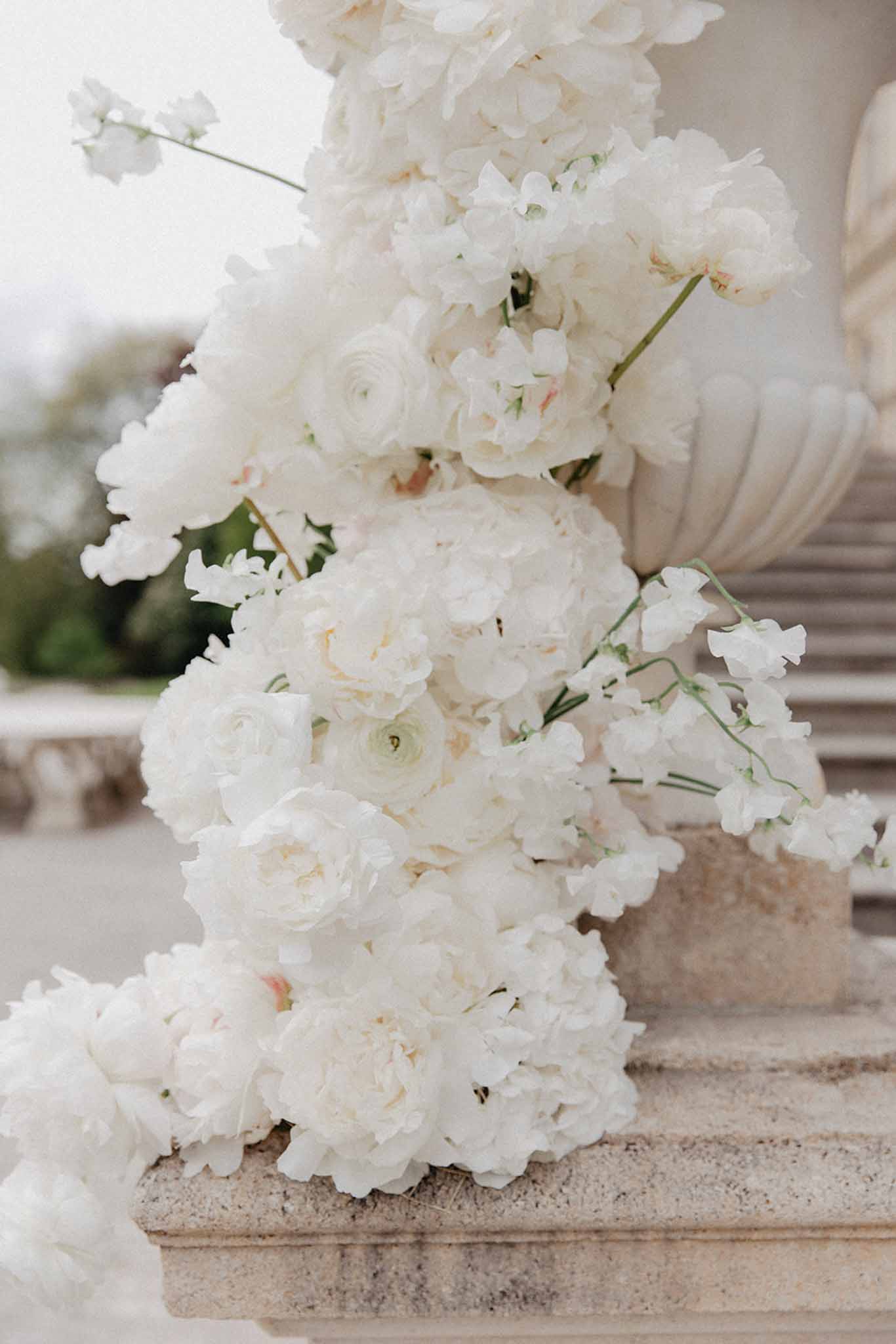 Close-up of ivory and cream peony bouquet with white sweet peas against stone balustrade with ornamental stonework in background