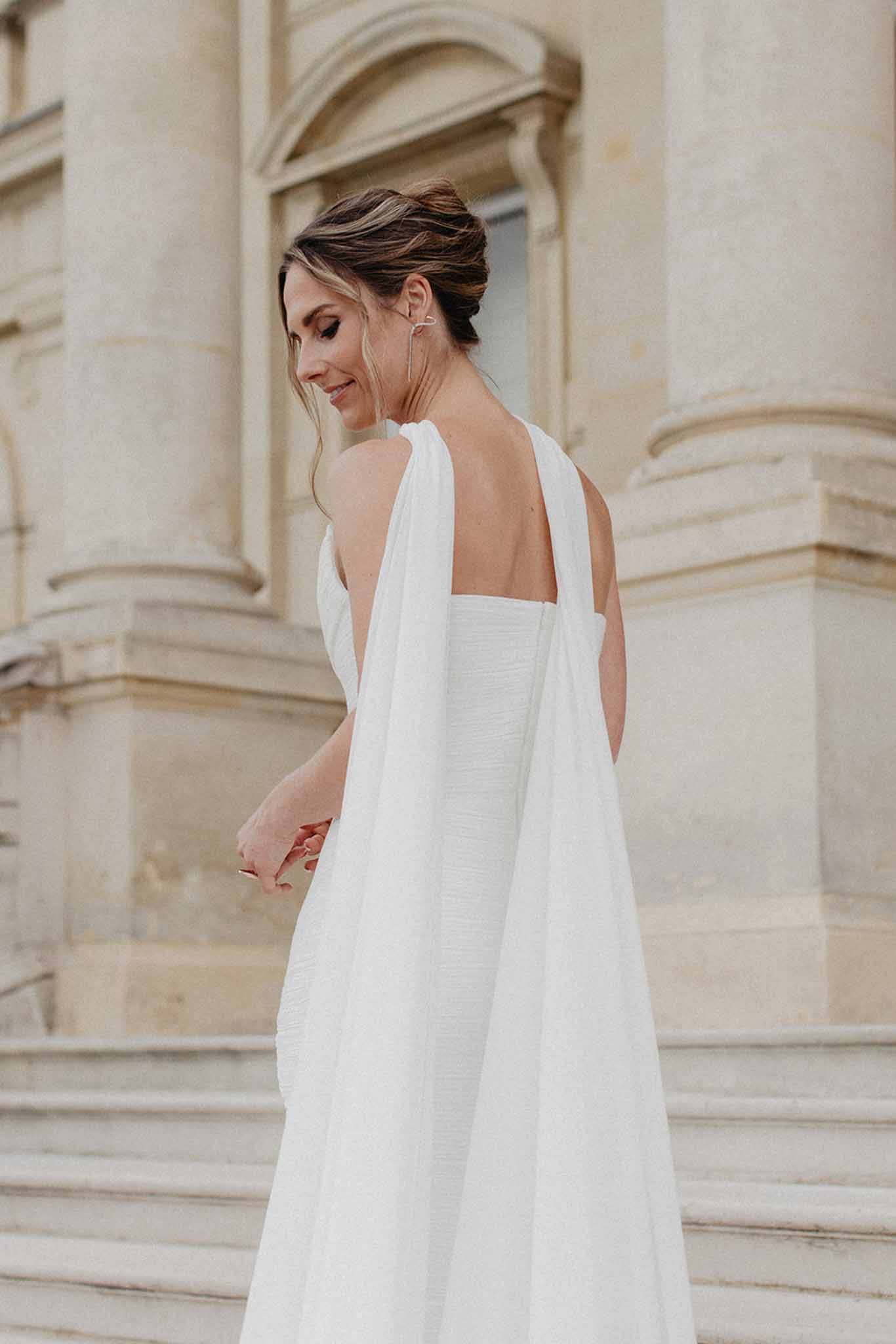 Bride in ivory A-line dress with cape detail posing in profile against classical stone building