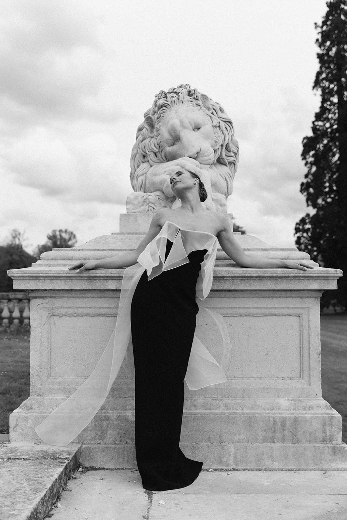 Bride in black strapless gown with white ruffled collar posing against stone lion sculpture in formal garden