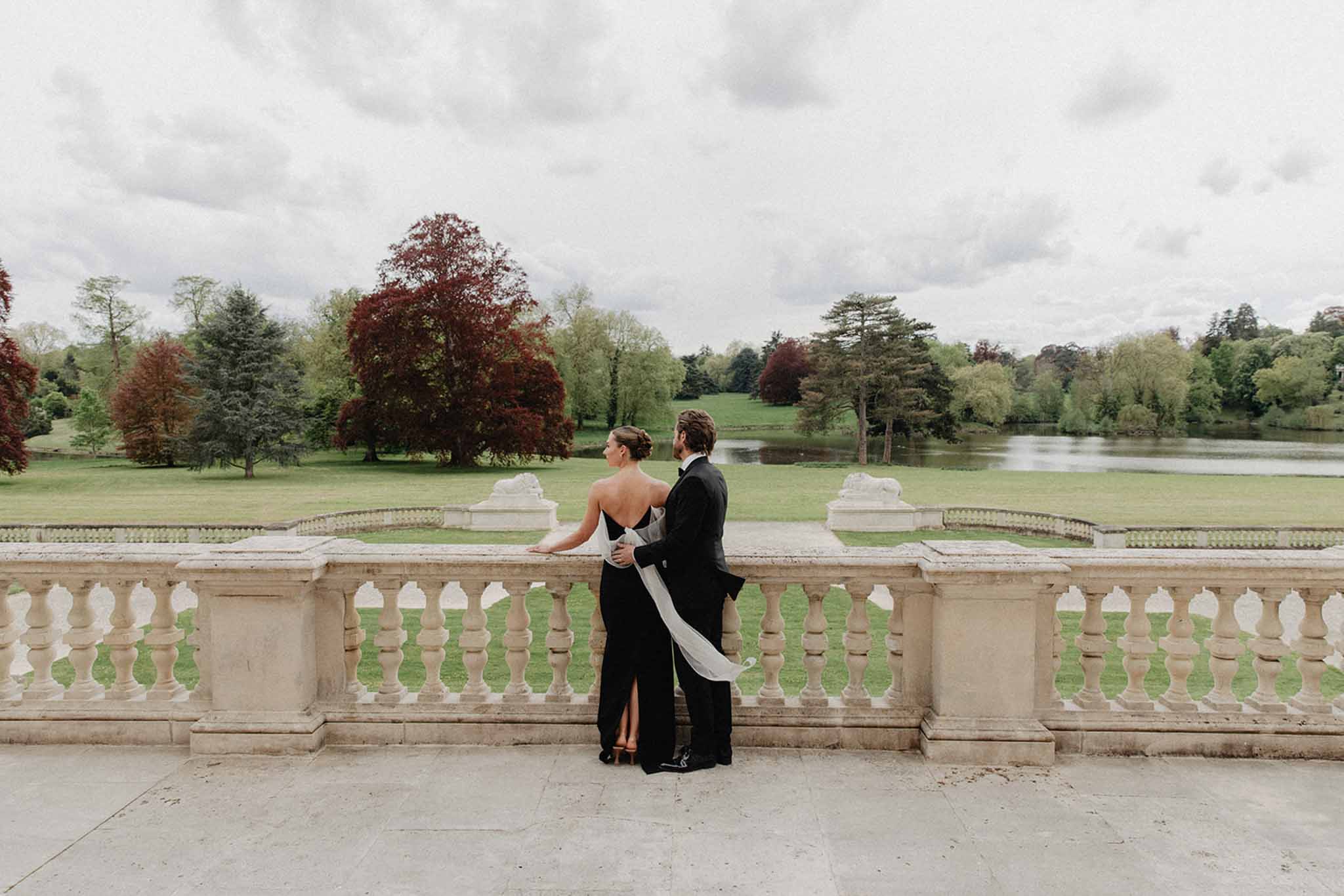 Bride and groom gazing over manicured grounds and lake from a stone terrace with classical balustrade