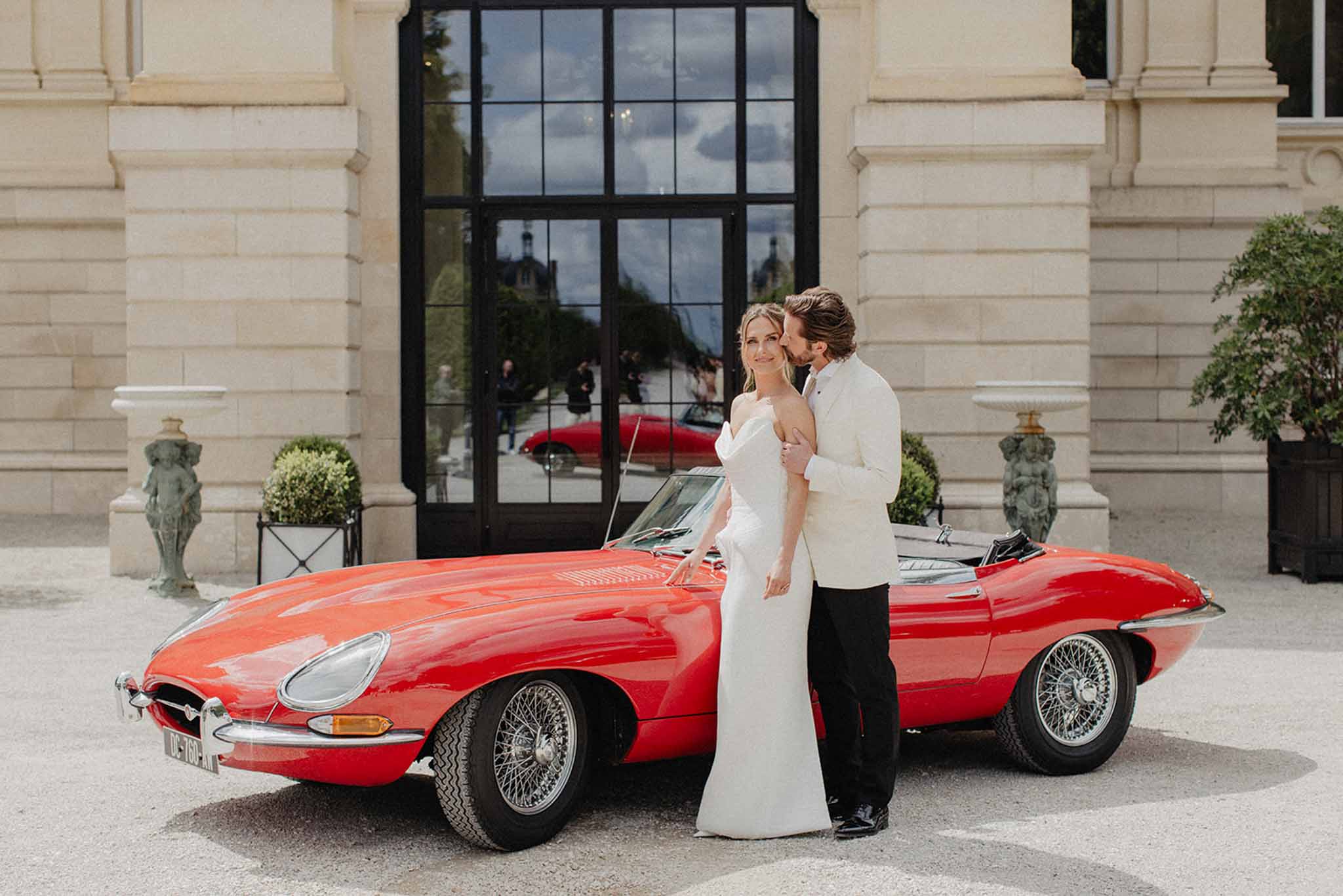 Bride and groom beside a vintage red sports car in a classical stone courtyard with potted boxwood topiaries.