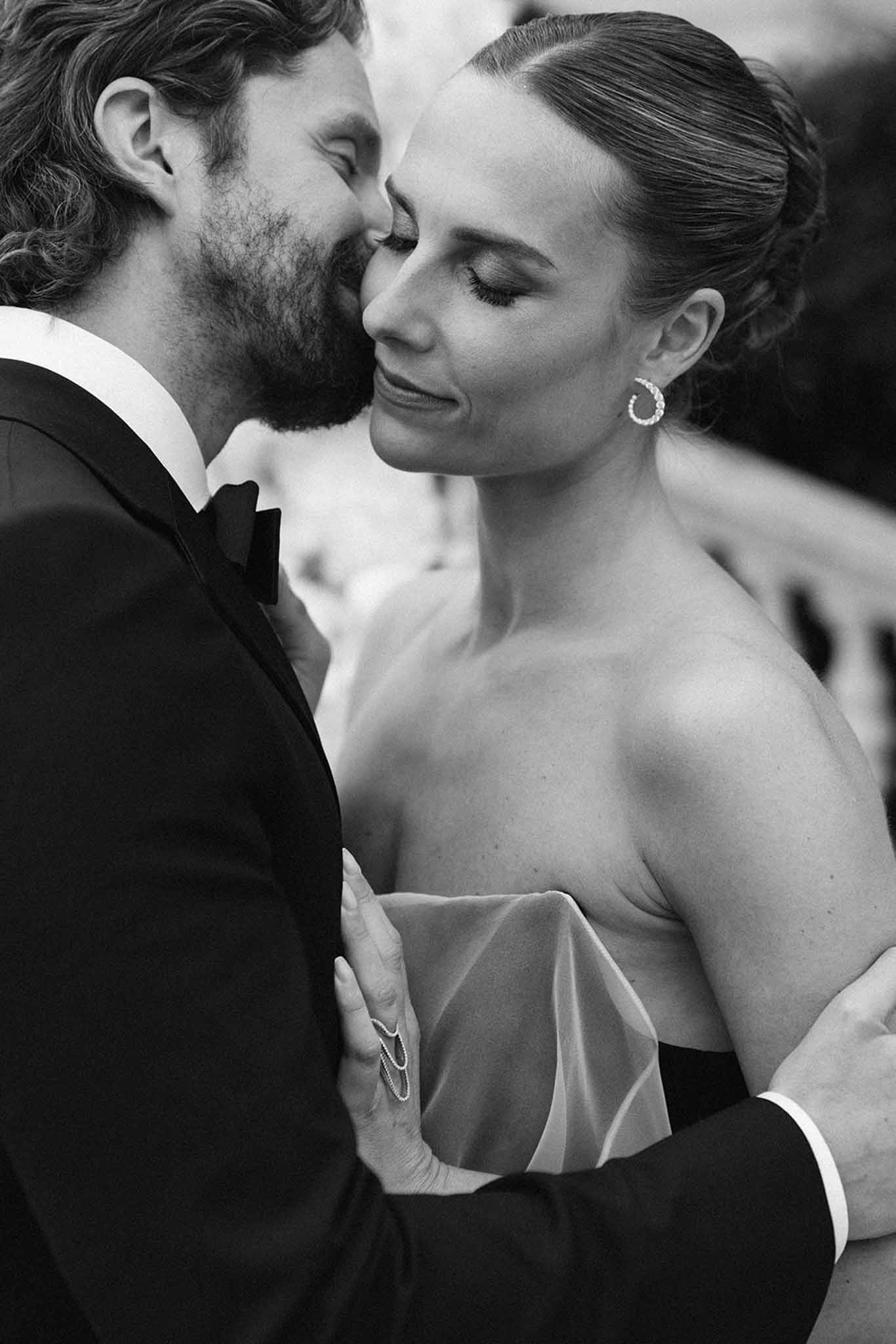 Black-and-white close-up of groom kissing bride's cheek, bride in strapless dress with hair pulled back