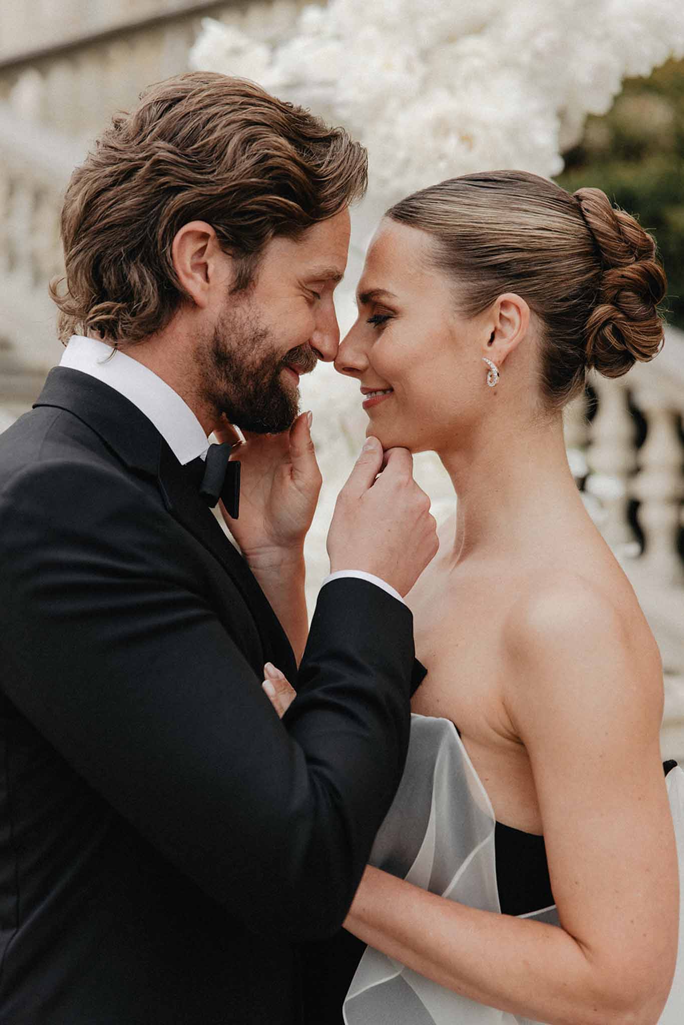 Bride and groom close together outdoors, foreheads almost touching; bride in ivory strapless gown, groom in black tuxedo.