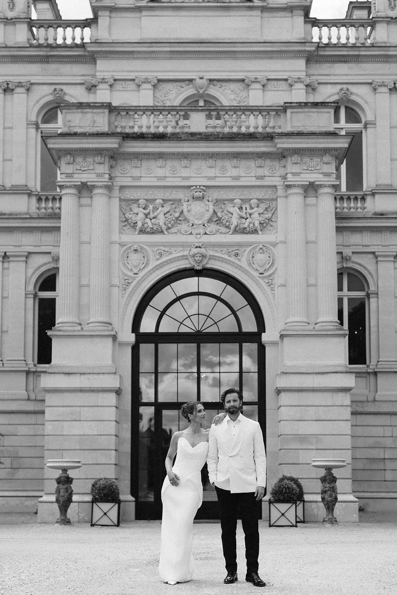 Black-and-white couple before grand neoclassical building with fluted columns, cherub reliefs, and boxwood topiary