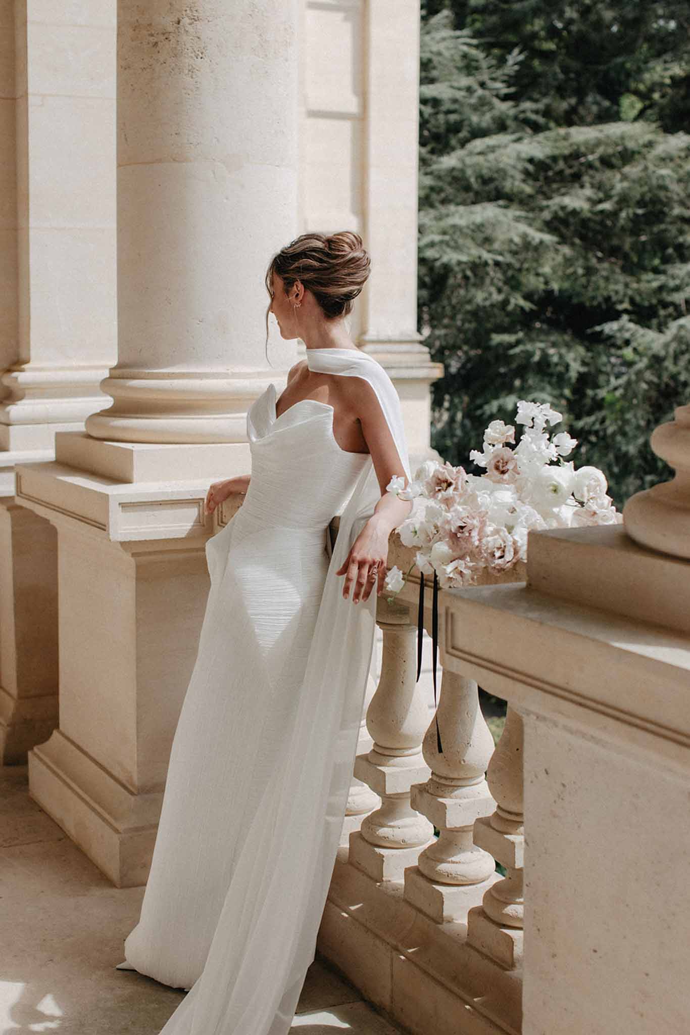 Bride in pleated halter gown holding peony bouquet on classical stone colonnade at Chateau de Ferrieres