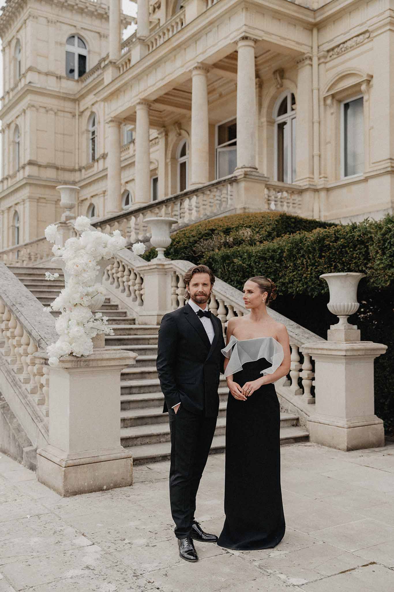 Bride in black gown and groom in tuxedo posing on stone courtyard of French mansion