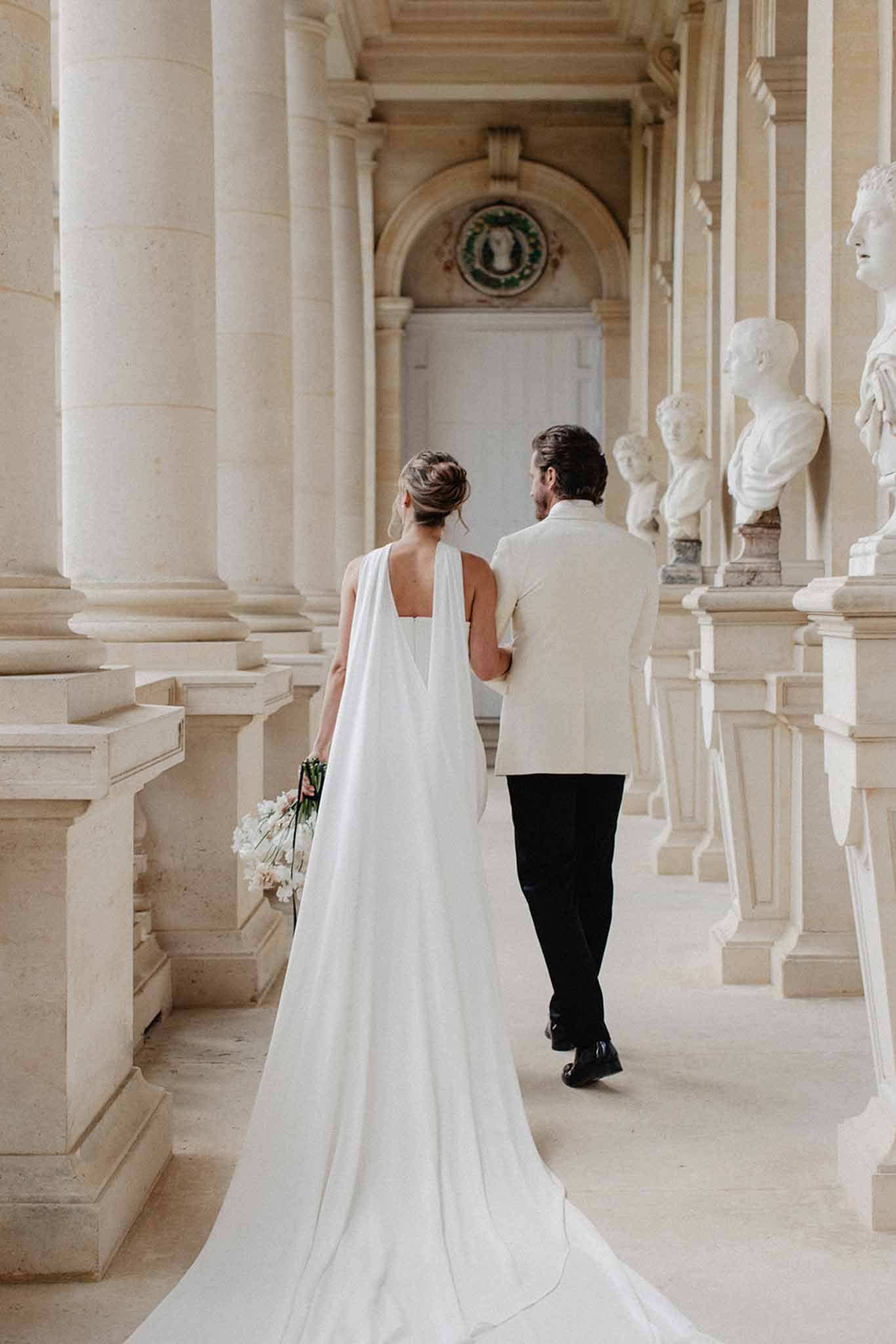 Couple walking through a neoclassical gallery lined with stone columns and white busts; bride in ivory gown with a flowing cape.