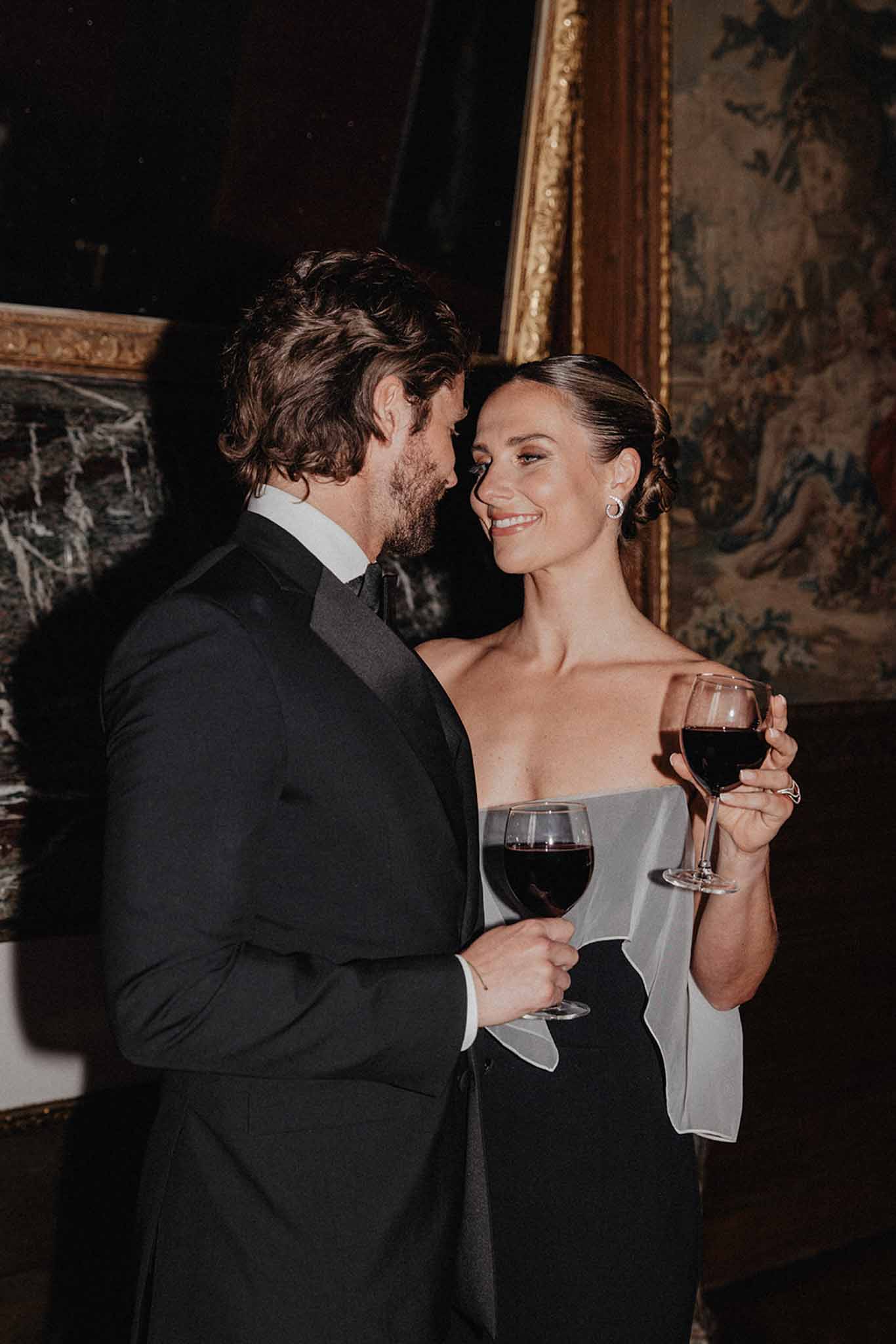 Bride in dove grey gown and groom in black tuxedo holding wine glasses during reception