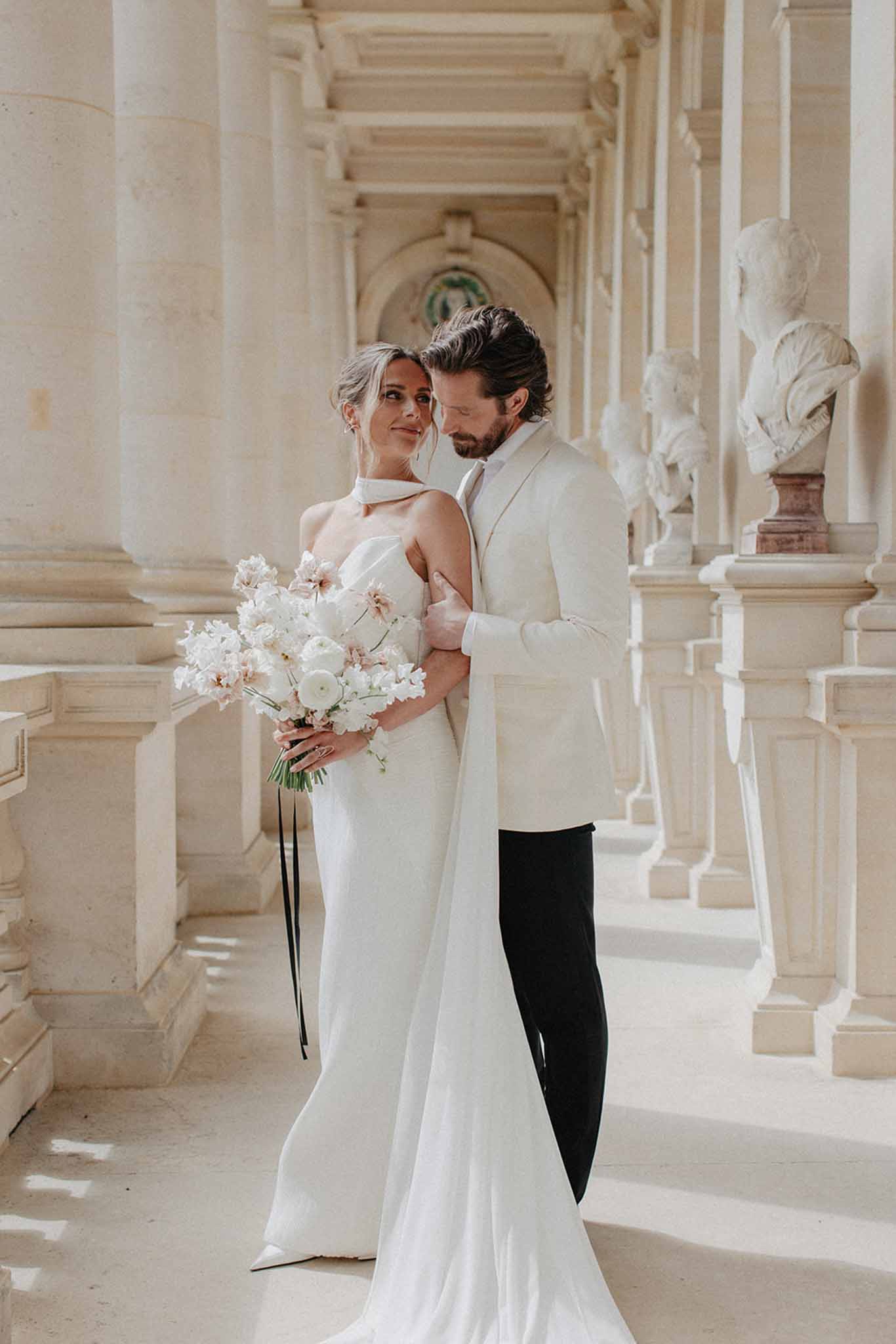 Bride in ivory halter column gown and groom in cream linen jacket posing in colonnaded corridor with marble busts