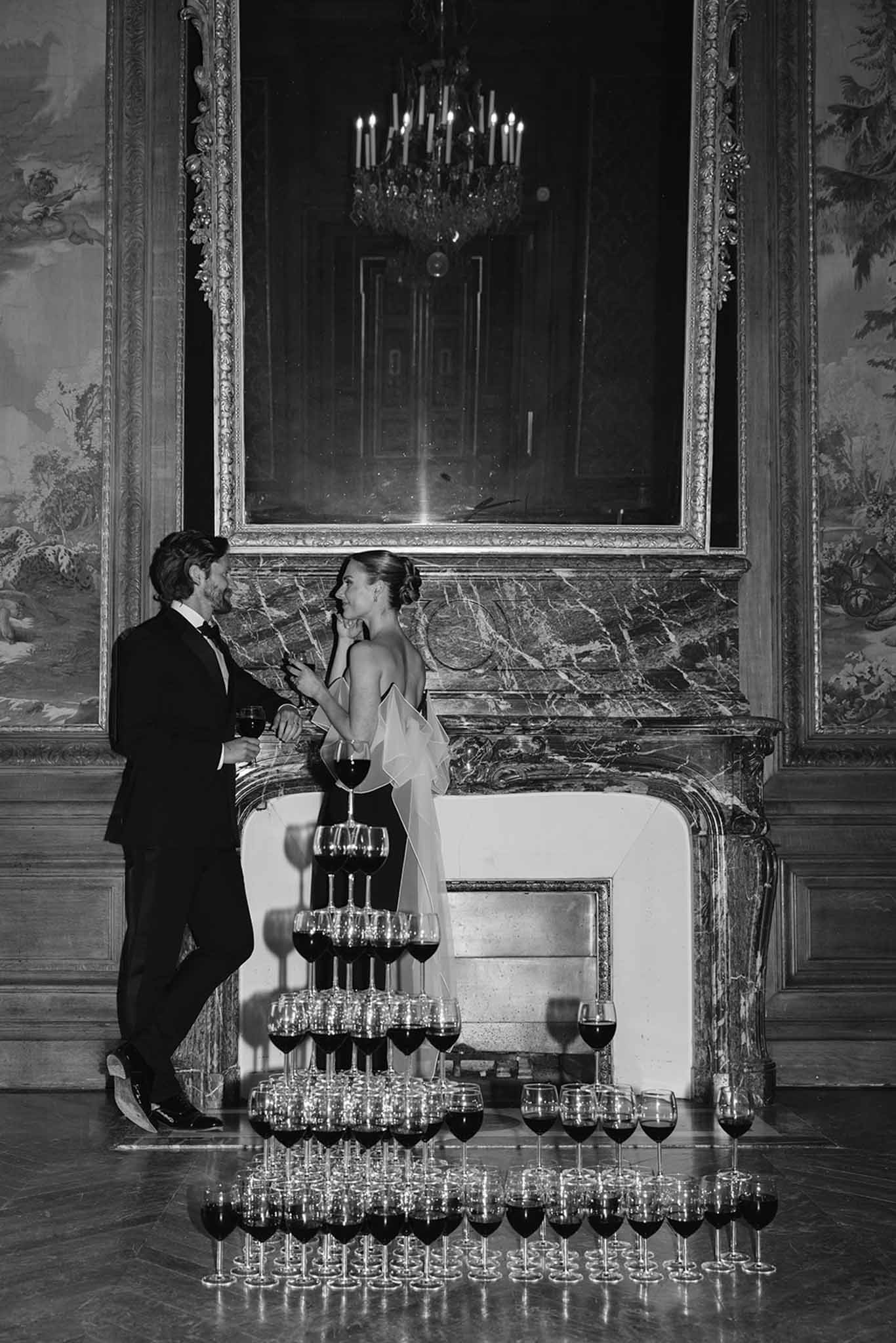 Black and white portrait of couple beside champagne coupe tower and ornate fireplace in palace interior