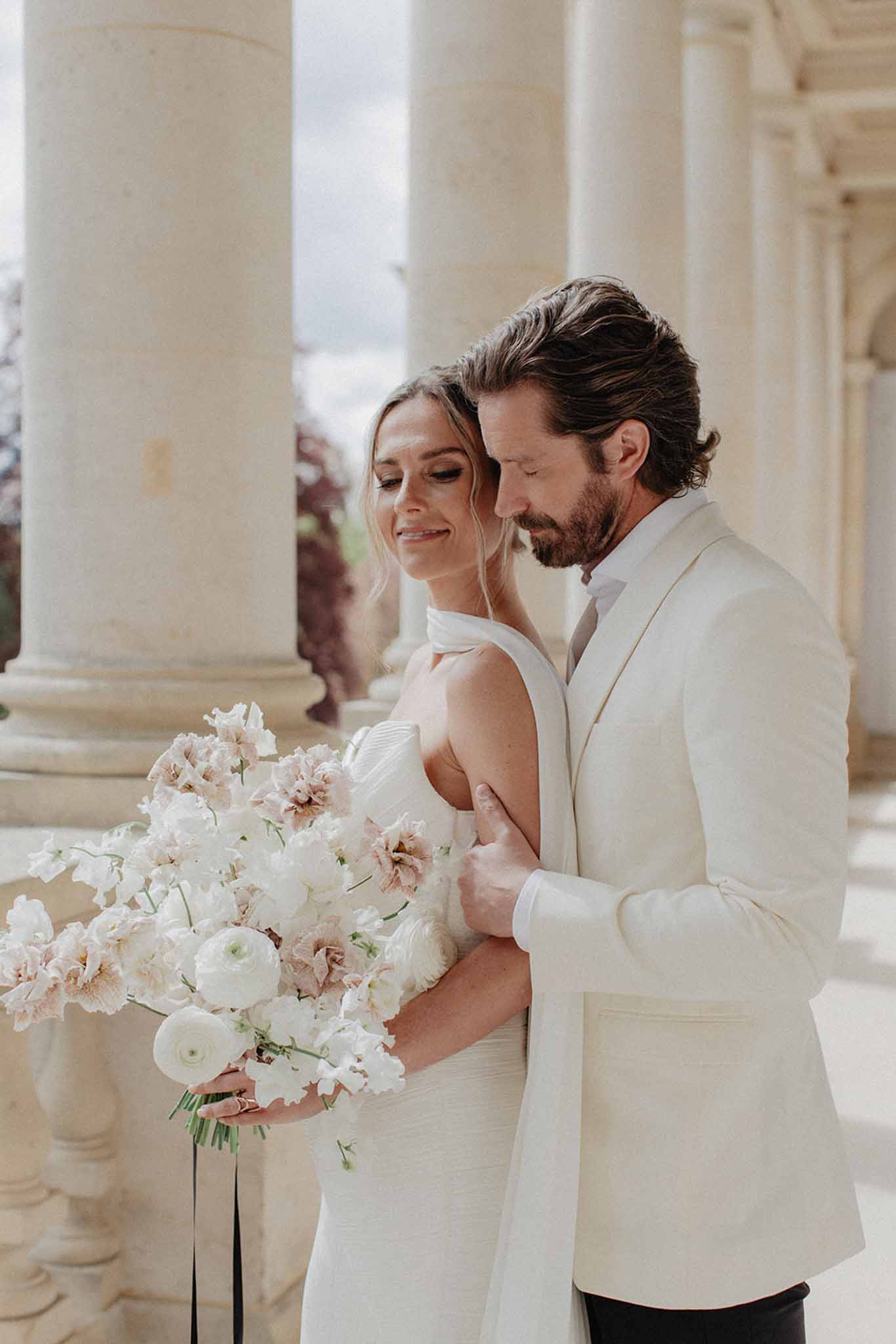 Bride and groom in columned corridor, bride holds white ranunculus and blush carnation bouquet, both in cream and ivory