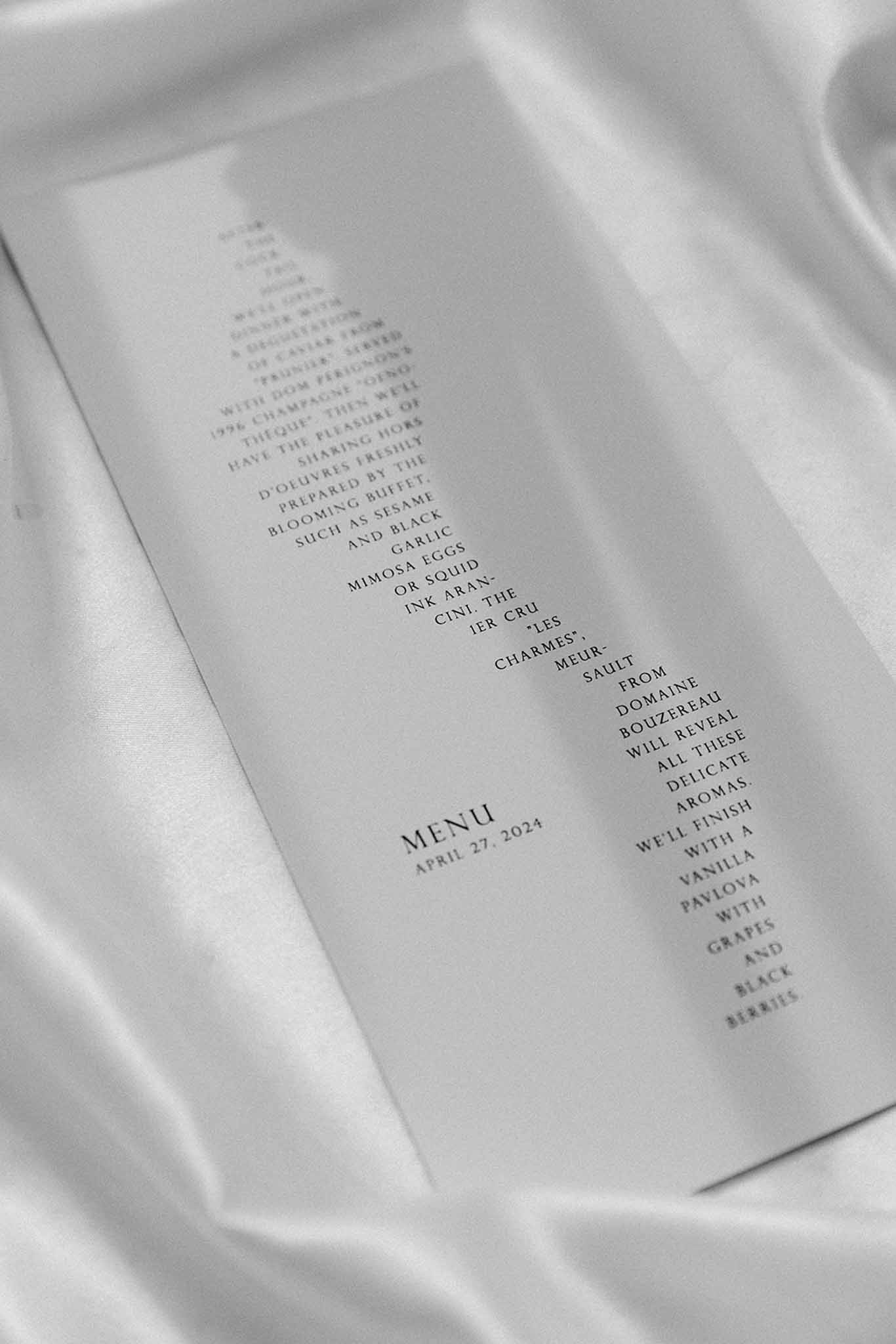 Black-and-white flat lay of multi-course wedding menu card on white silk fabric dated April 2024