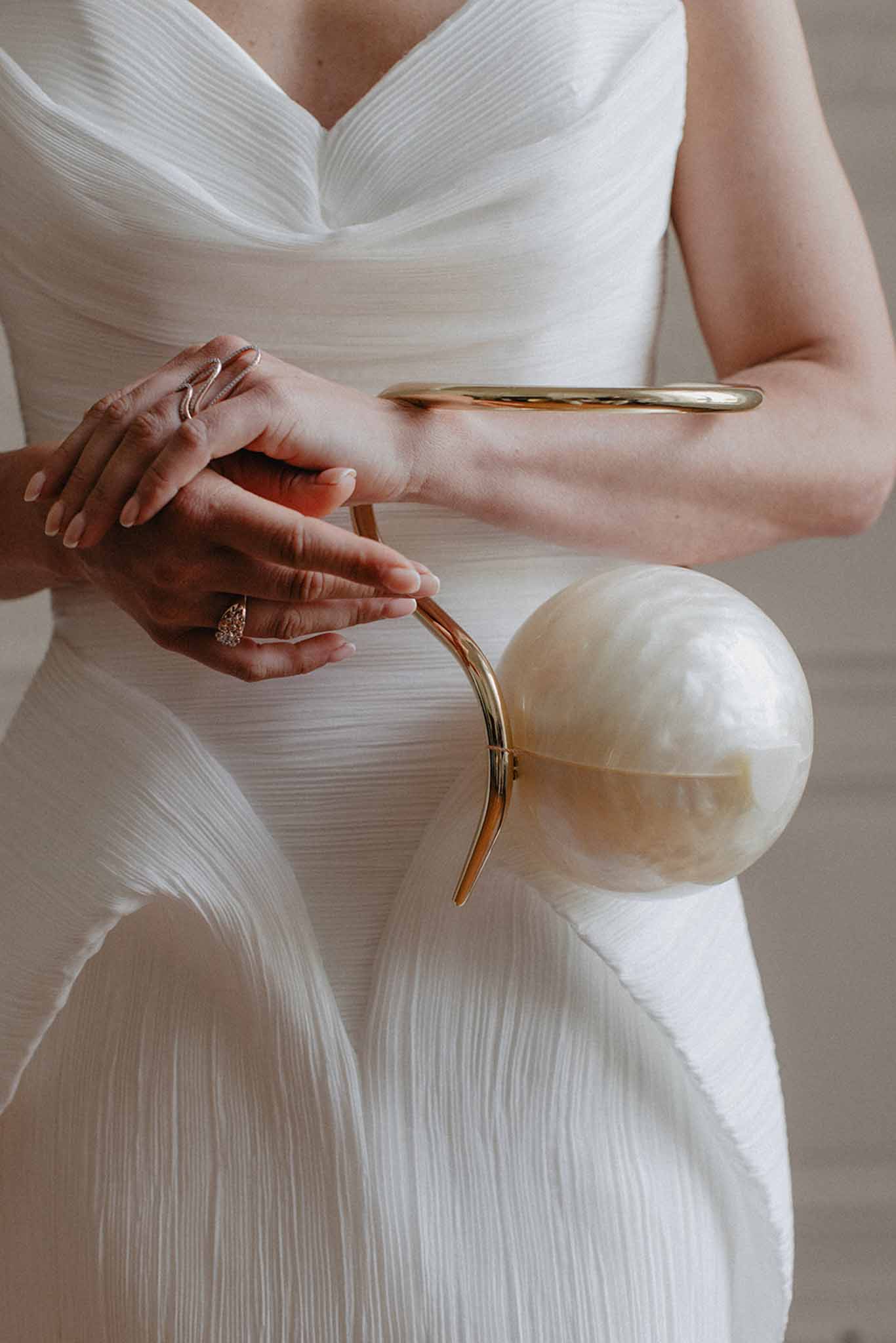 Bride in pleated cowl-neck ivory dress holds clear acrylic clutch with gold handle, diamond ring visible