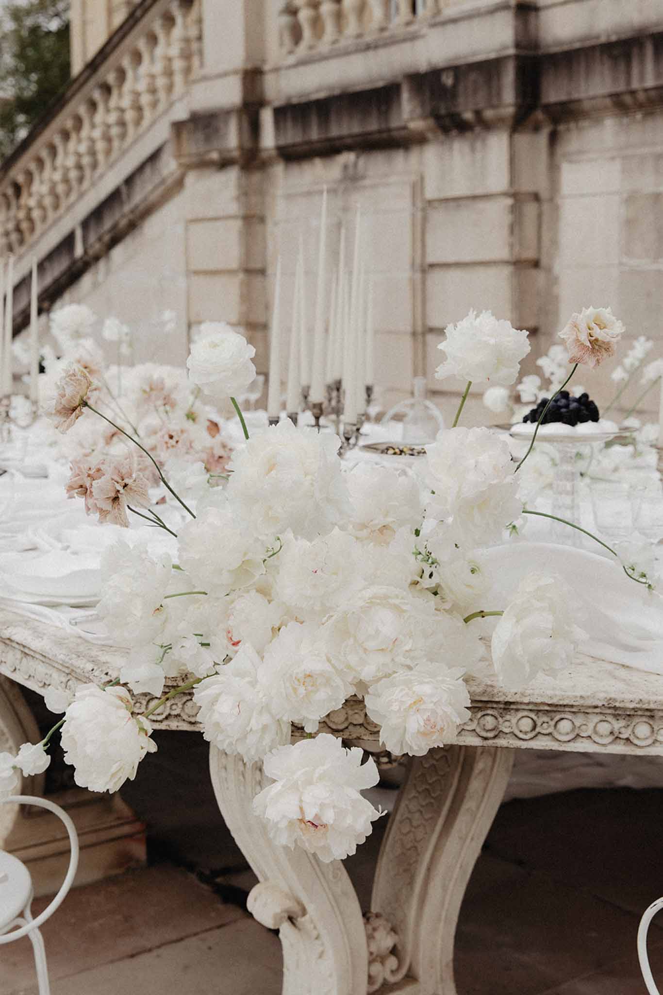 White ornamental urn centerpiece with ivory and blush peonies and roses on carved stone pedestal at outdoor reception table