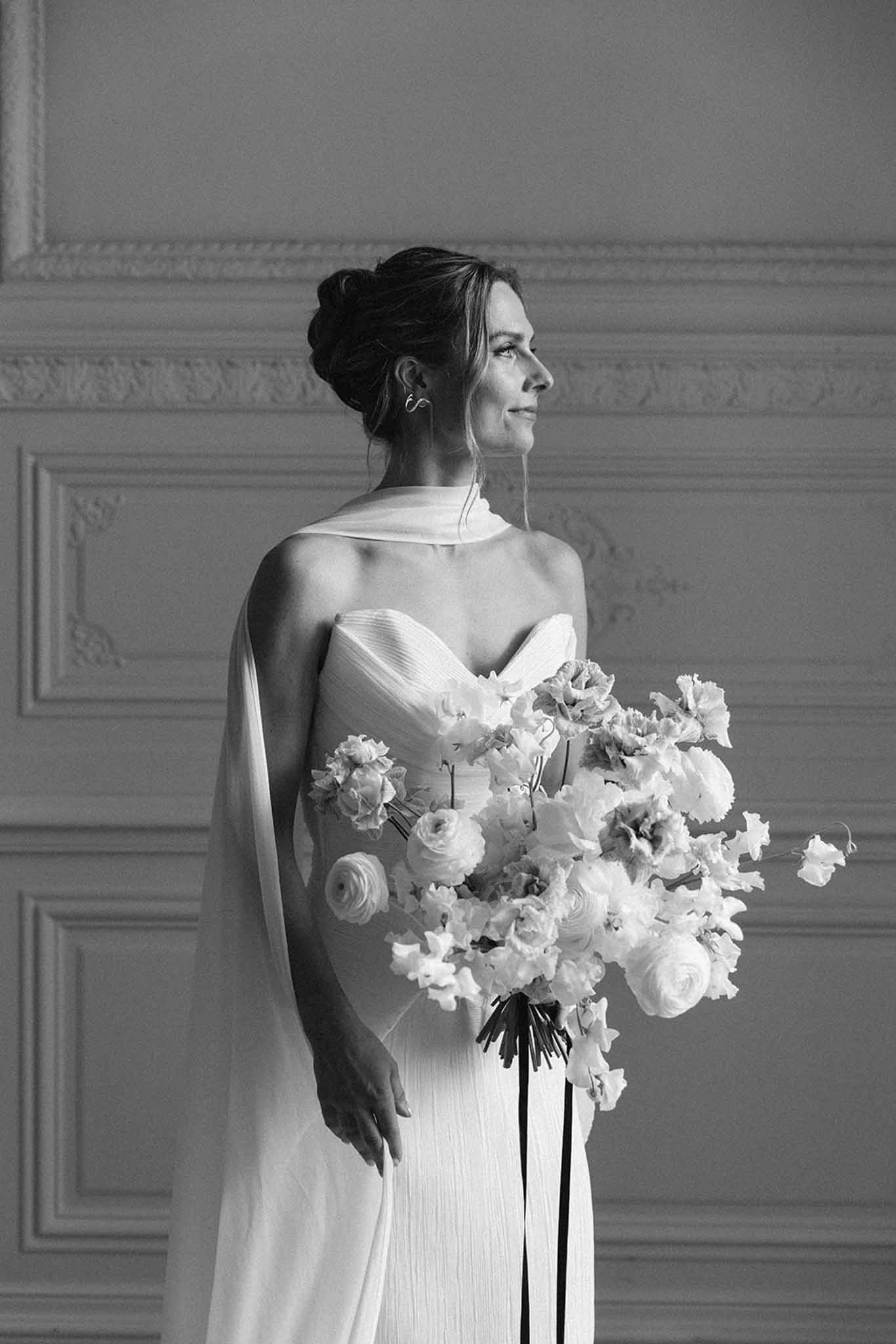 Black and white bridal portrait showing pleated gown with cape and large bouquet in classical interior