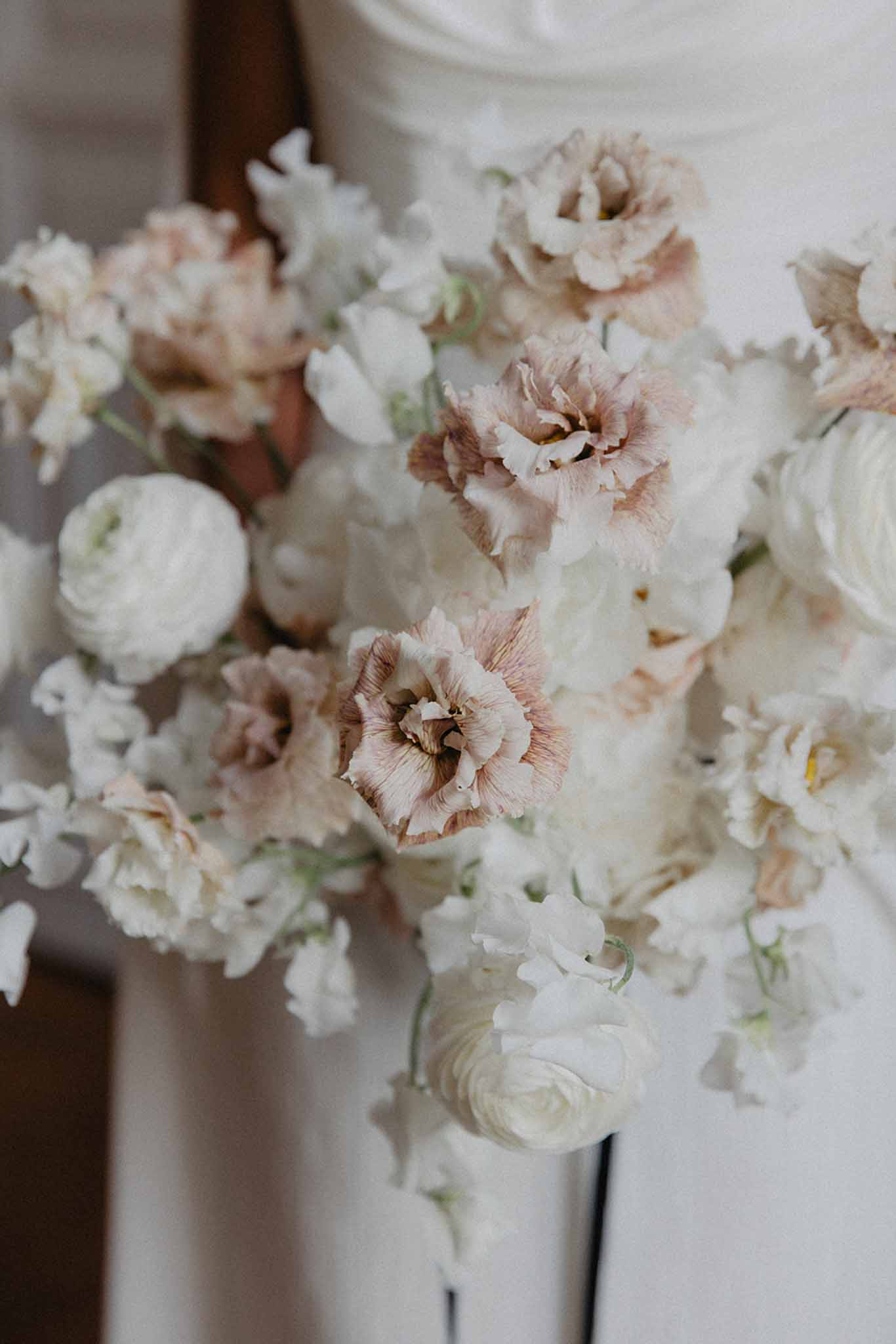 Close-up of bridal bouquet with dusty rose carnations, ivory roses and white ranunculus against white wedding dress