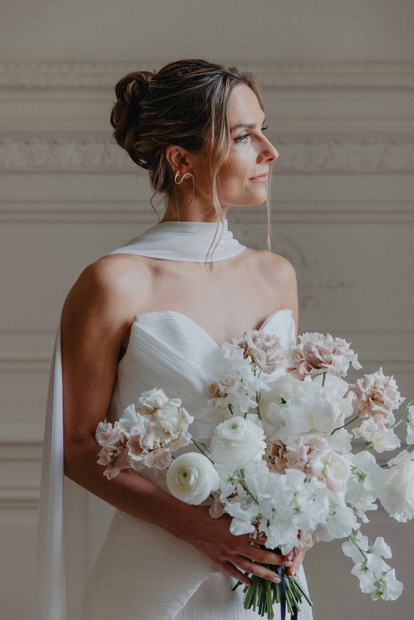 Side profile bridal portrait with ivory ranunculus and blush pink bouquet against beige wall