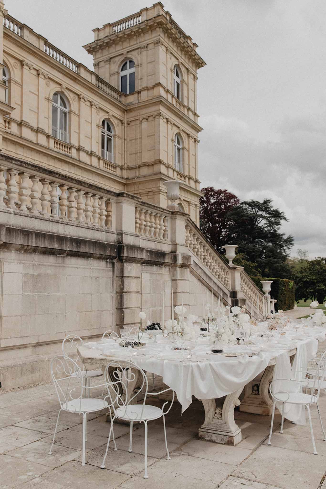 Outdoor reception on stone terrace in front of grand neoclassical mansion with long ivory-draped tables and white chairs