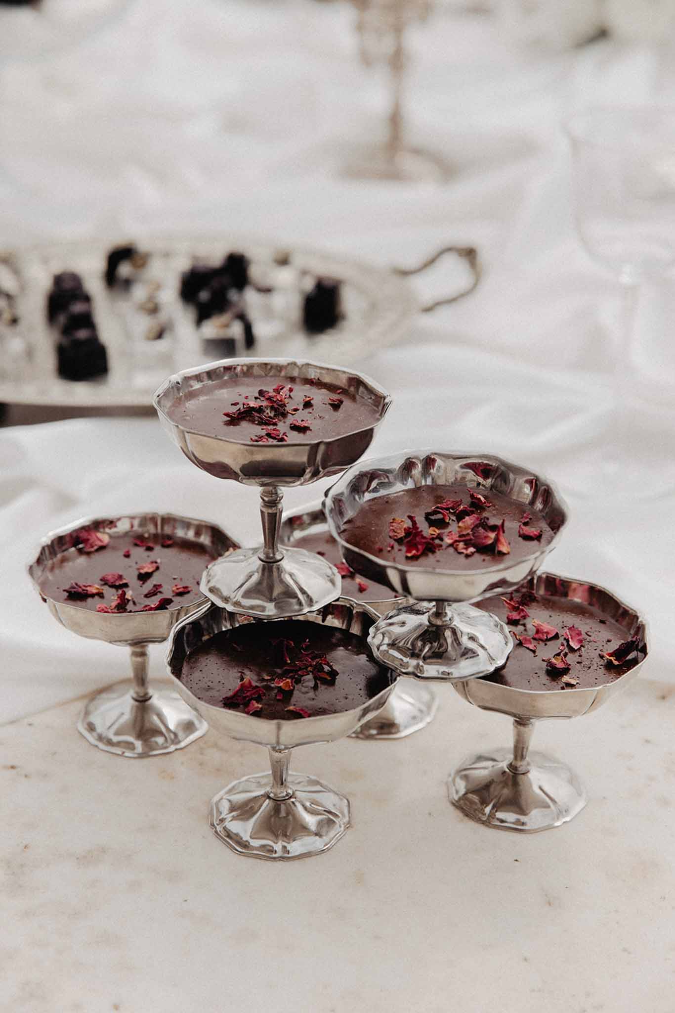 Five silver coupe glasses filled with dark chocolate mousse topped with dried rose petals at reception