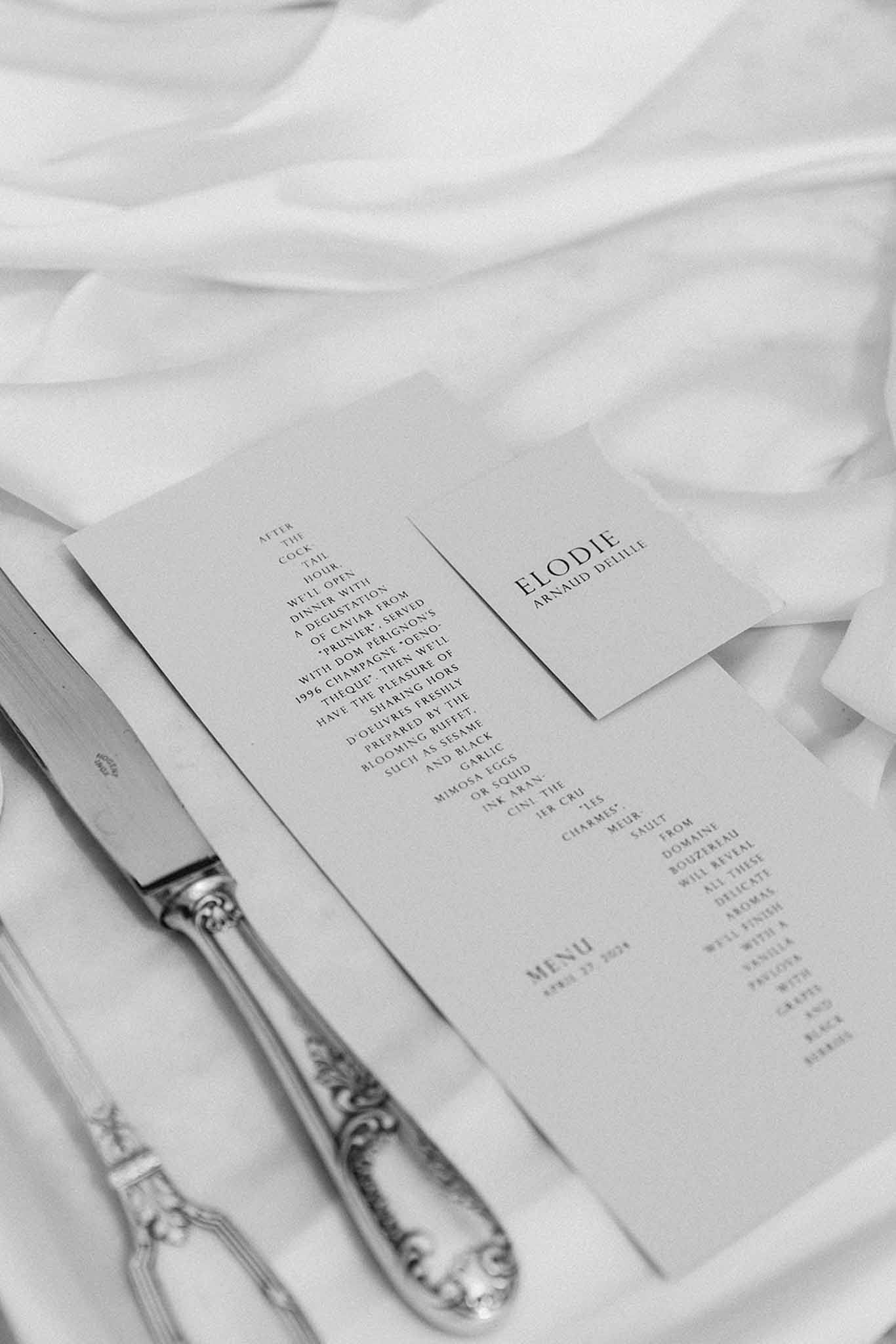 Black-and-white flat lay of wedding reception cards and couple names on white fabric alongside ornate silverware