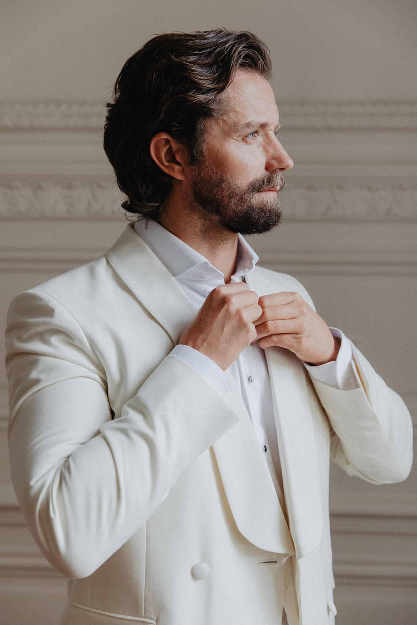 Groom in cream double-breasted suit adjusting his jacket in profile against neutral taupe paneled interior wall