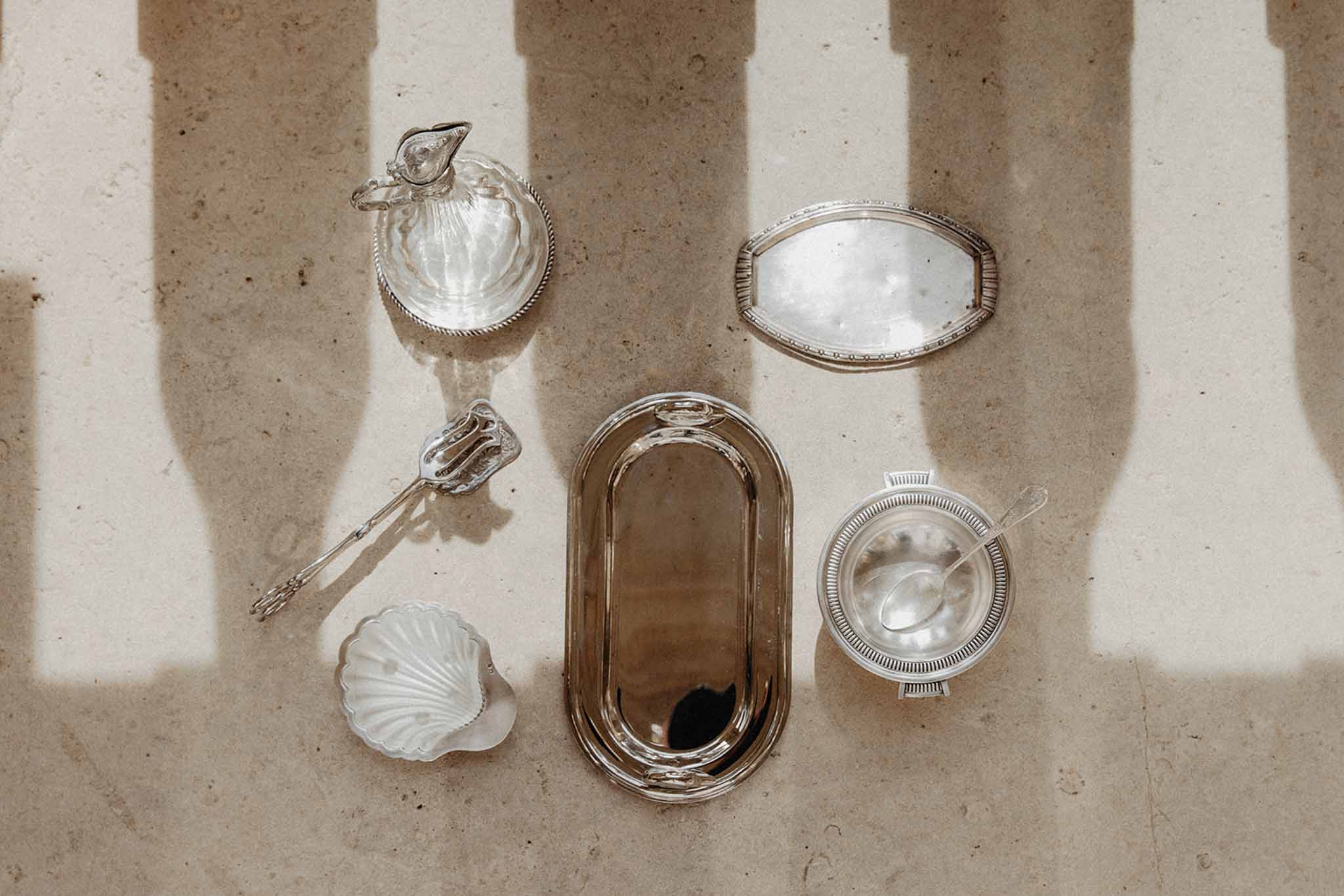 Flat lay of six antique silver serving pieces including pitcher, trays and shell dish arranged on stone surface