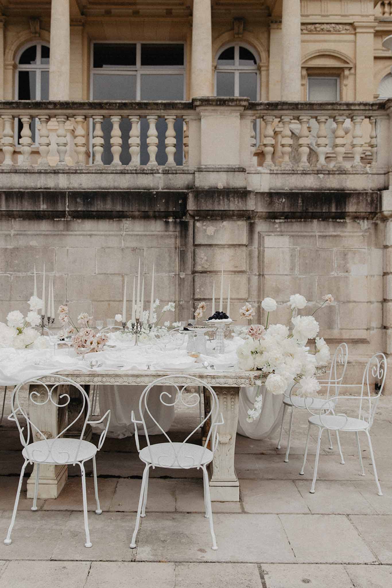 Outdoor terrace reception table with white peonies and dahlias, taper candles, and ornate metal chairs