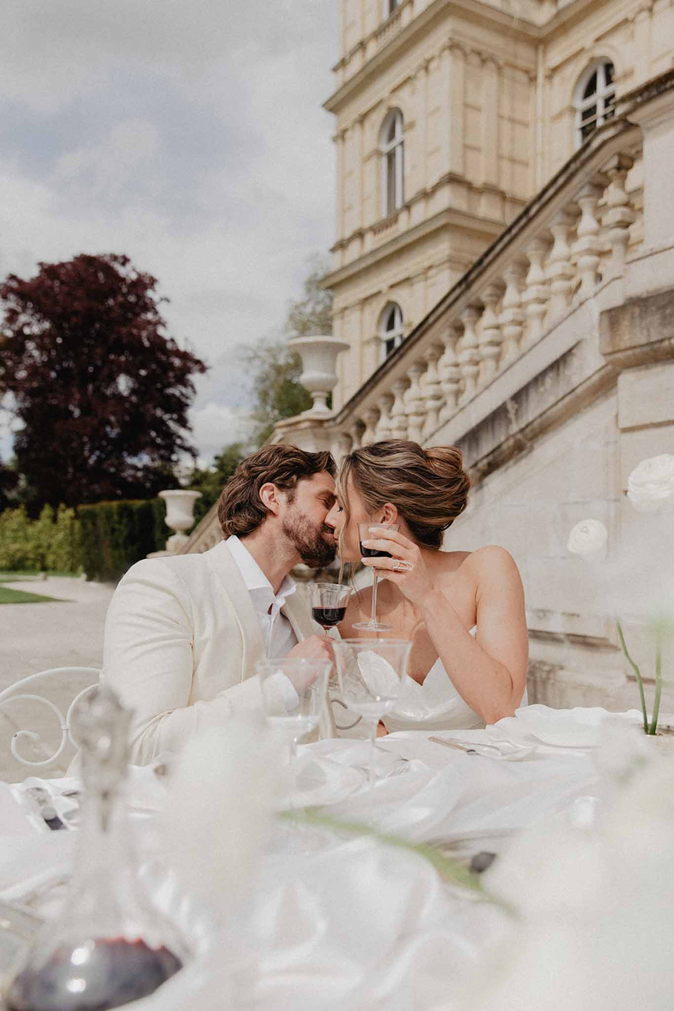 Bride and groom touching foreheads at outdoor reception table against cream stone mansion, both holding wine glasses