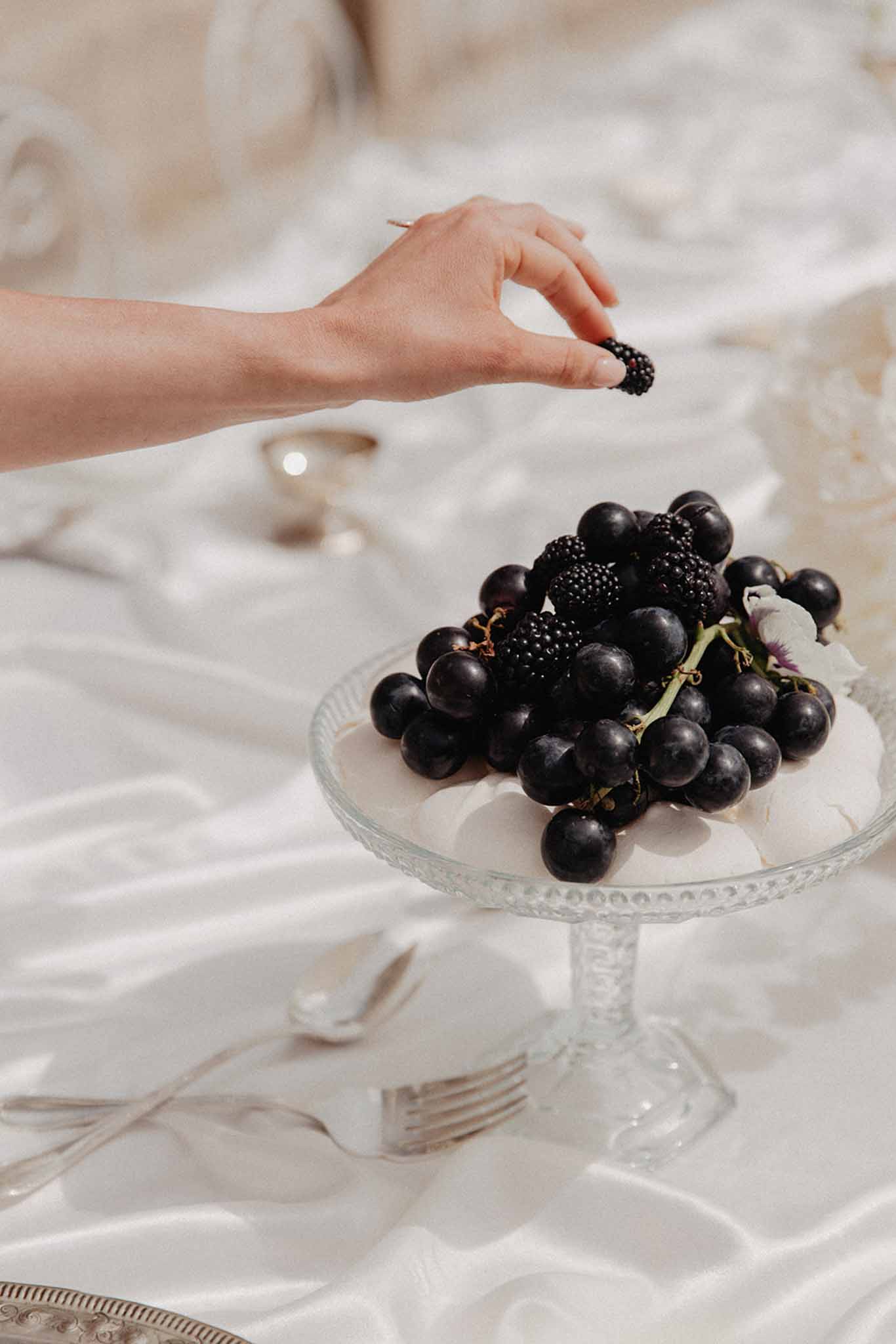 Glass pedestal dish of black grapes and blackberries on a cream linen reception table with white plates and cutlery.