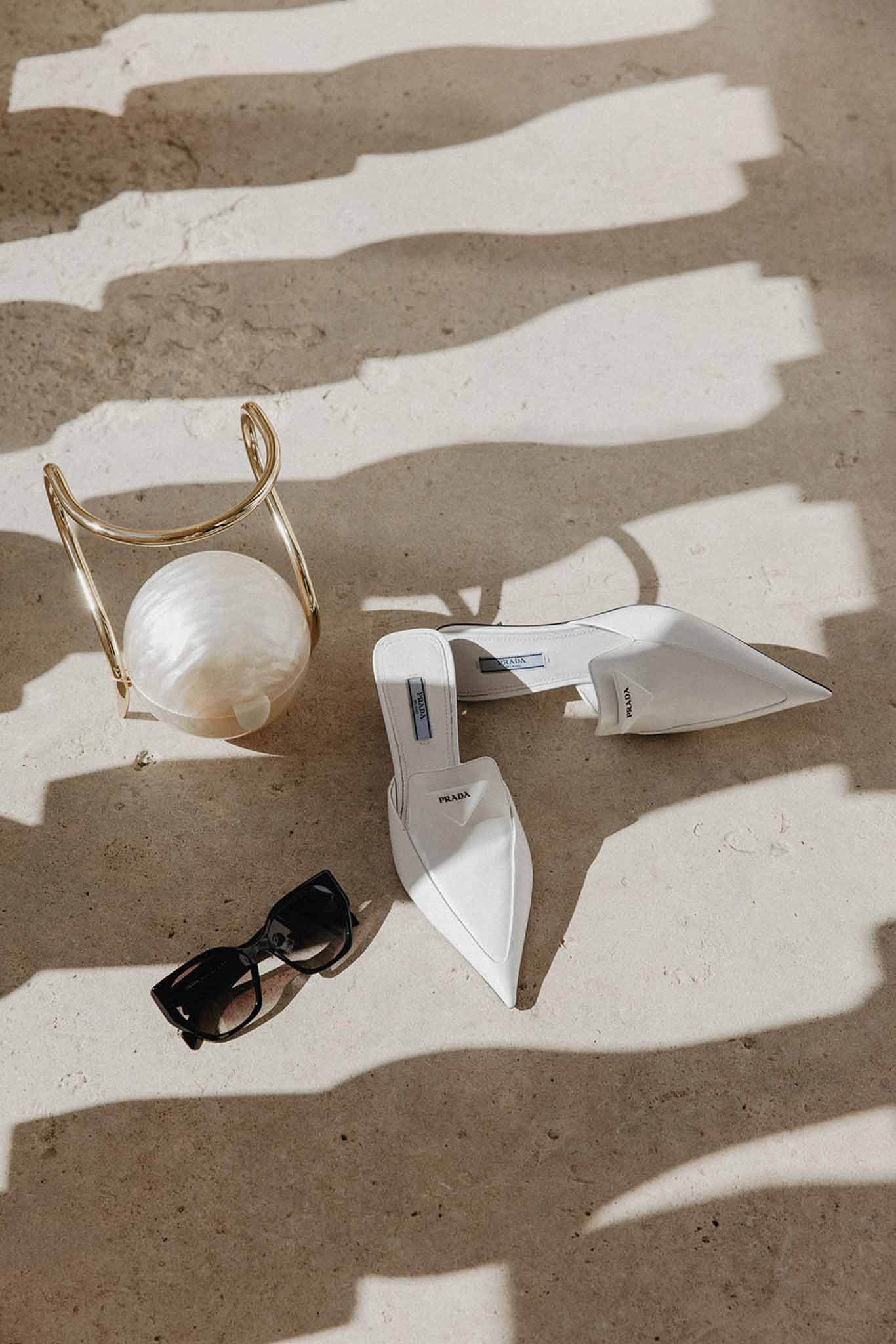 Flat lay of bridal accessories with white Prada mules, clear clutch, and sunglasses on concrete with striped shadows