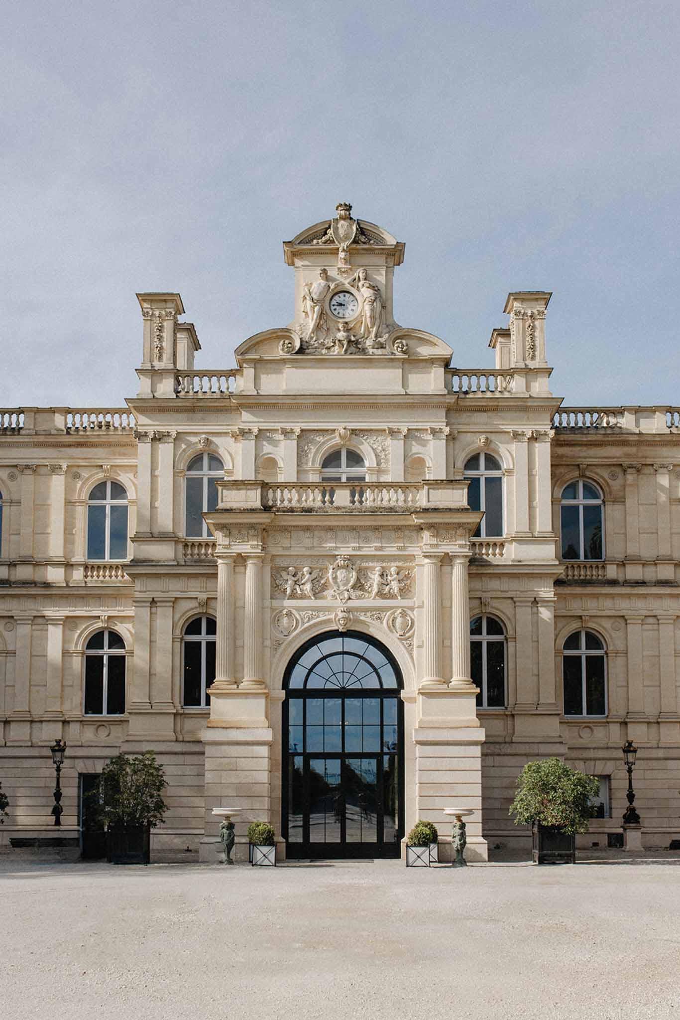 Neoclassical stone chateau facade with arched windows, fluted columns, clock tower, and courtyard approach