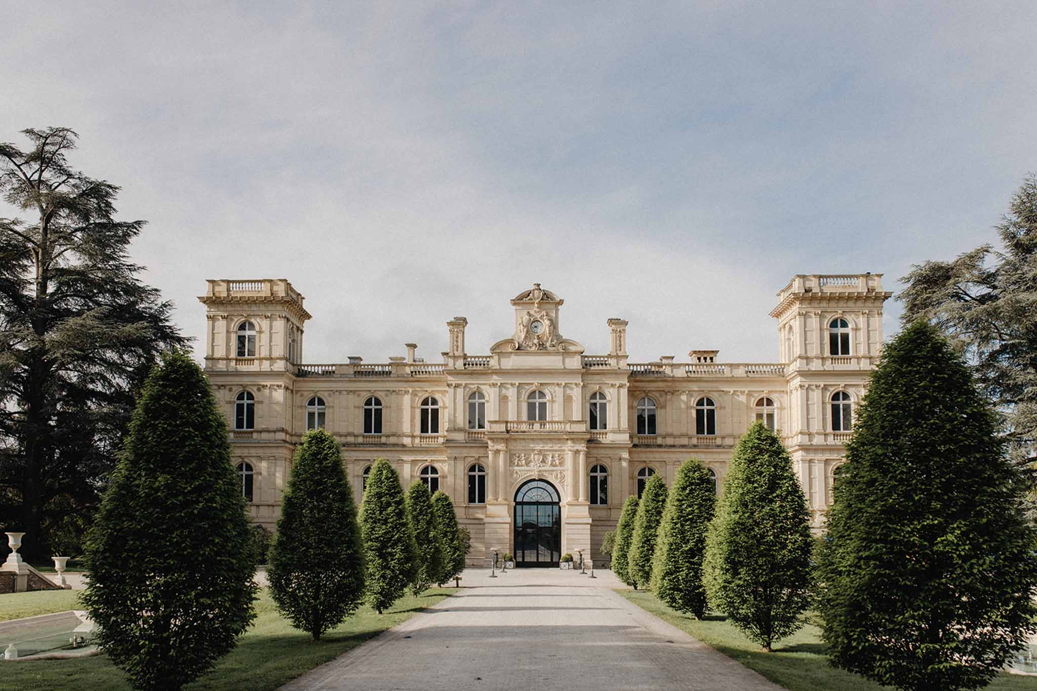 Symmetrical cream stone chateau facade with mansard roofs and conical topiaries lining the main approach