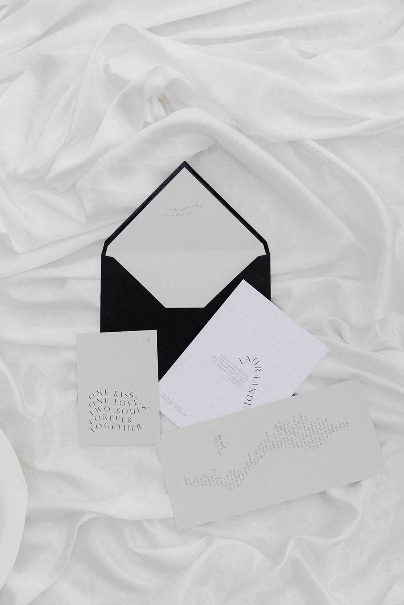 Flat lay of minimalist wedding stationery with black envelope and cream invitation cards on white tulle