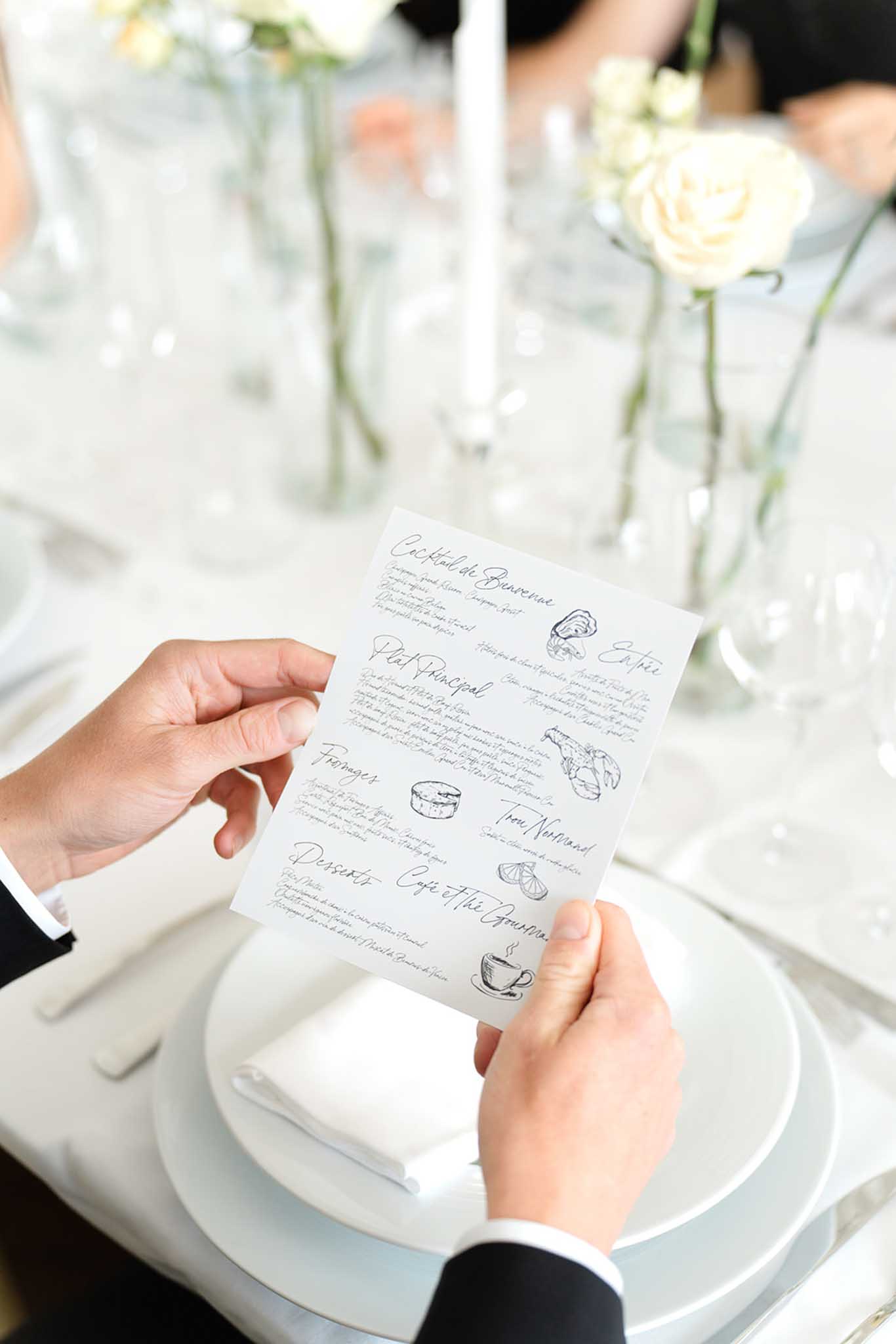 Close-up of groom's hands holding handwritten menu card at white place setting with cream roses in glass vases in background